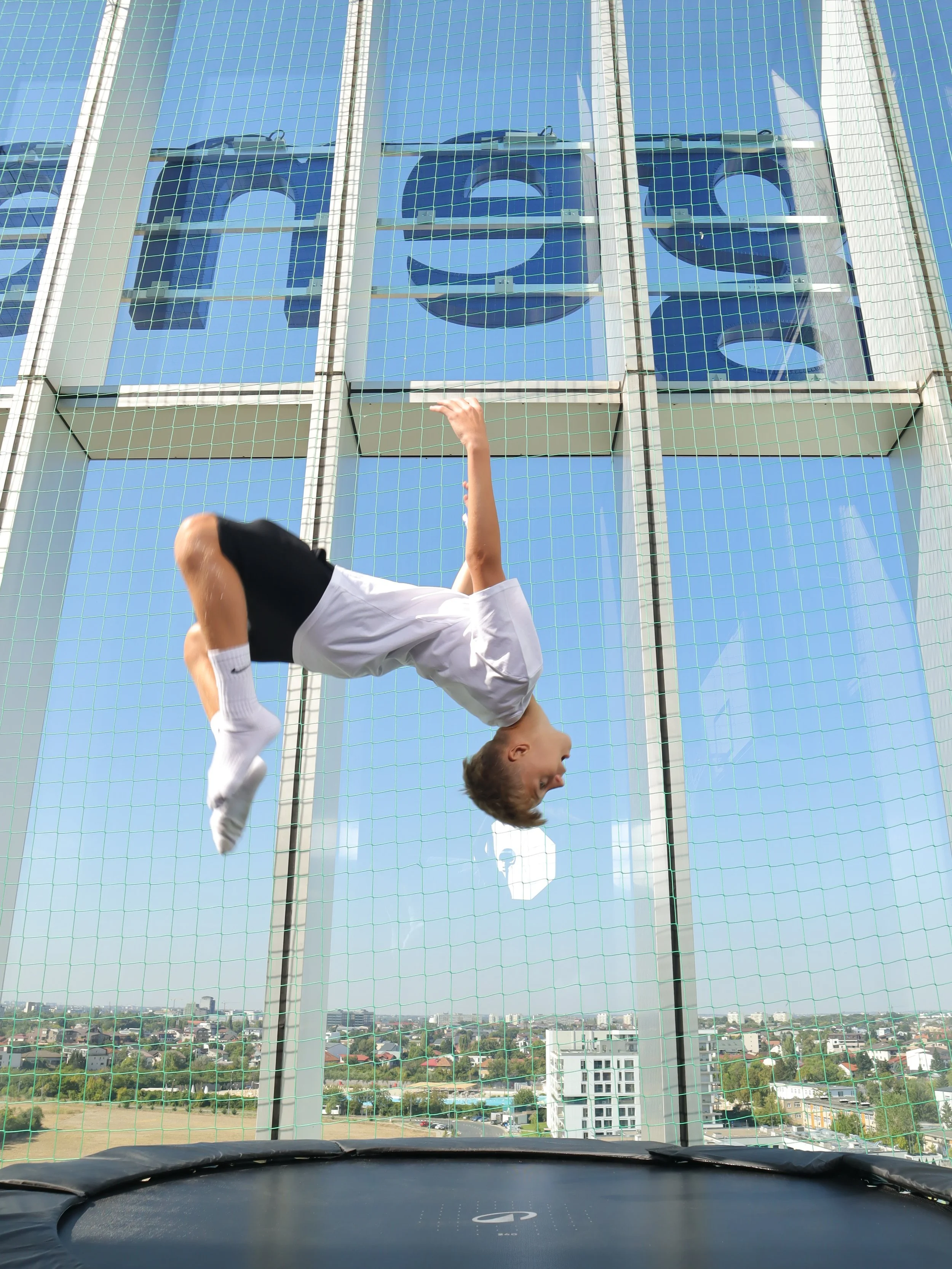 View from inside a car looking at a young boy falling upside down onto a trampoline through a safety net, with a cityscape and blue sky in the background.