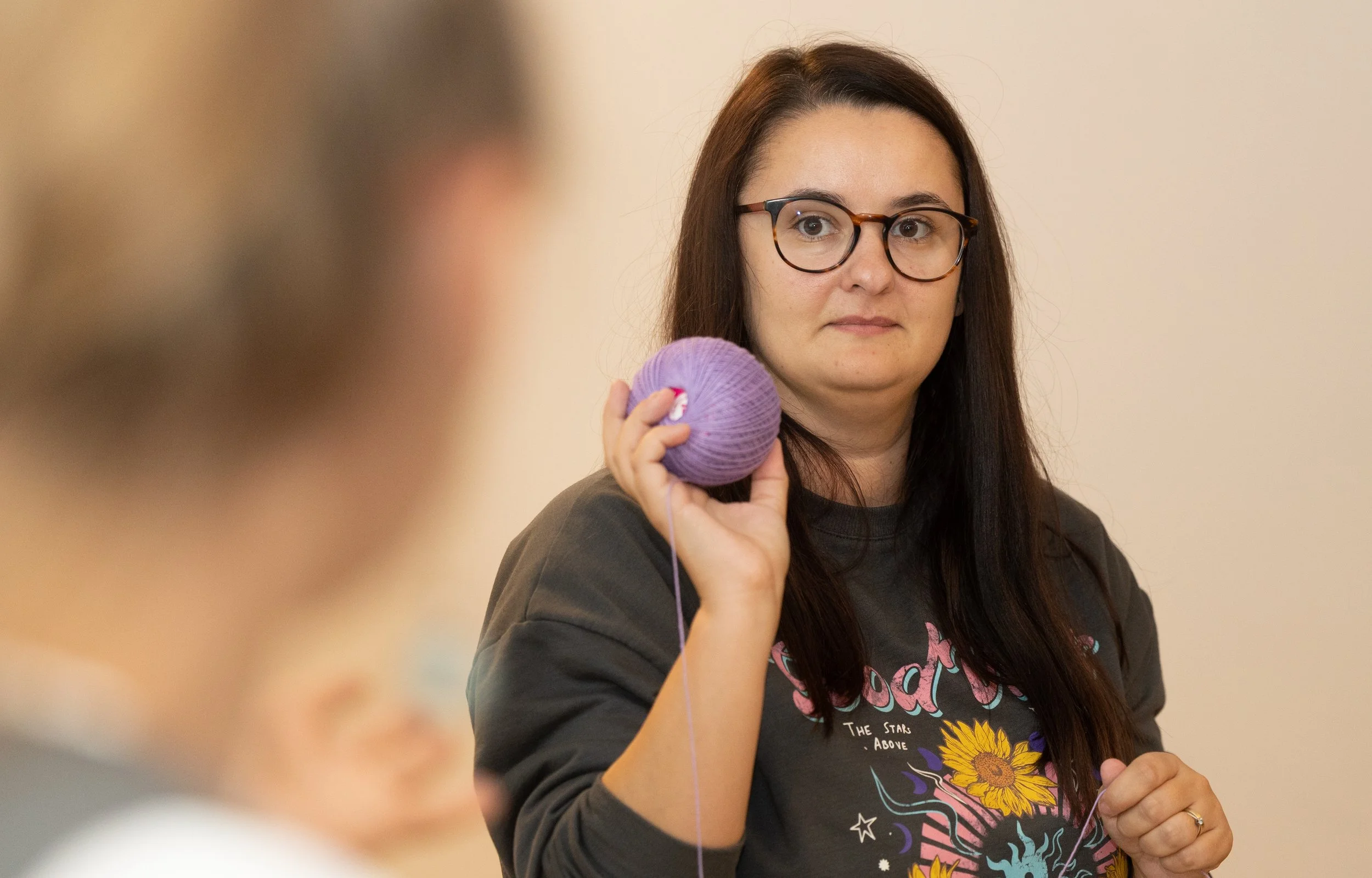 A woman with long dark hair and glasses holding purple yarn at knitting or craft class, wearing a dark sweatshirt with colorful graphic design.