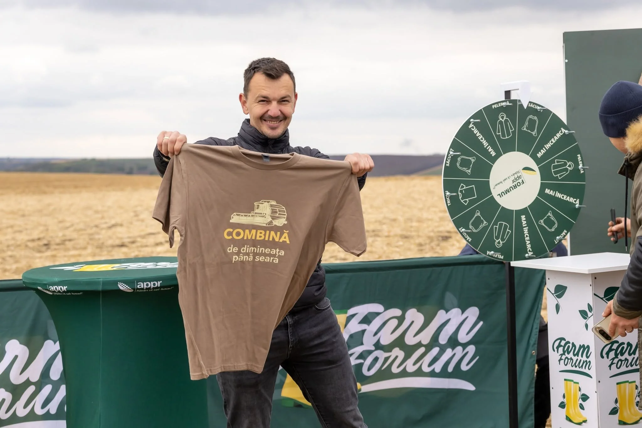 A man smiling and holding a brown T-shirt with a yellow graphic of a combine harvester and Romanian text, standing outdoors at a farm forum event. There is a green table and a wheel of fortune with icons and Romanian text nearby.