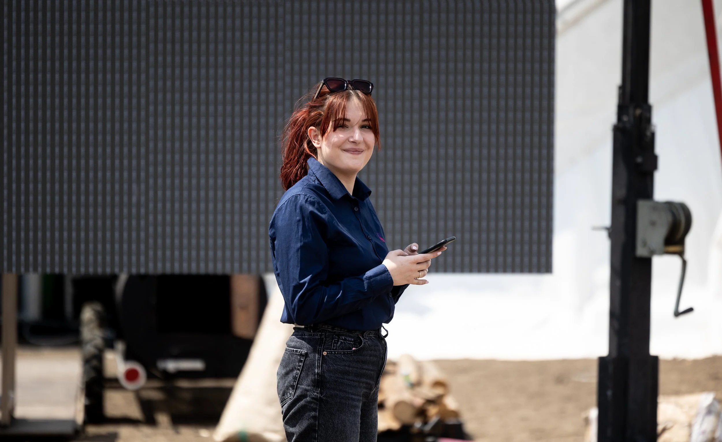 A woman with red hair wearing a navy blue shirt and dark jeans, holding a phone, standing outdoors near large solar panels.