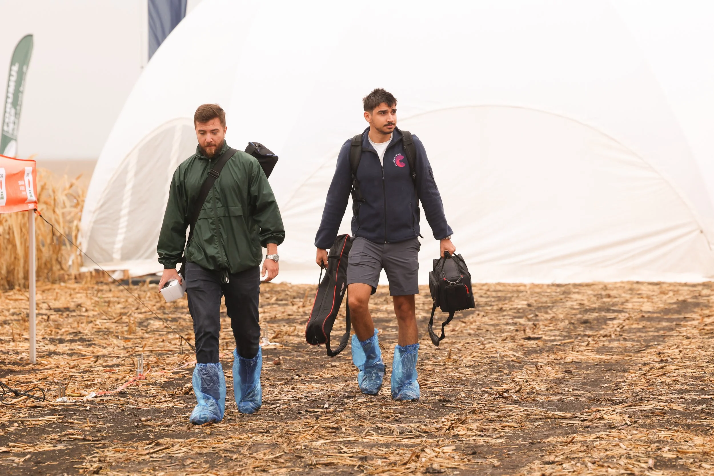 Two young men walking through a field with ashes and debris, wearing blue protective shoe covers, carrying backpacks, and holding equipment, with a large white inflatable structure and some banners in the background.