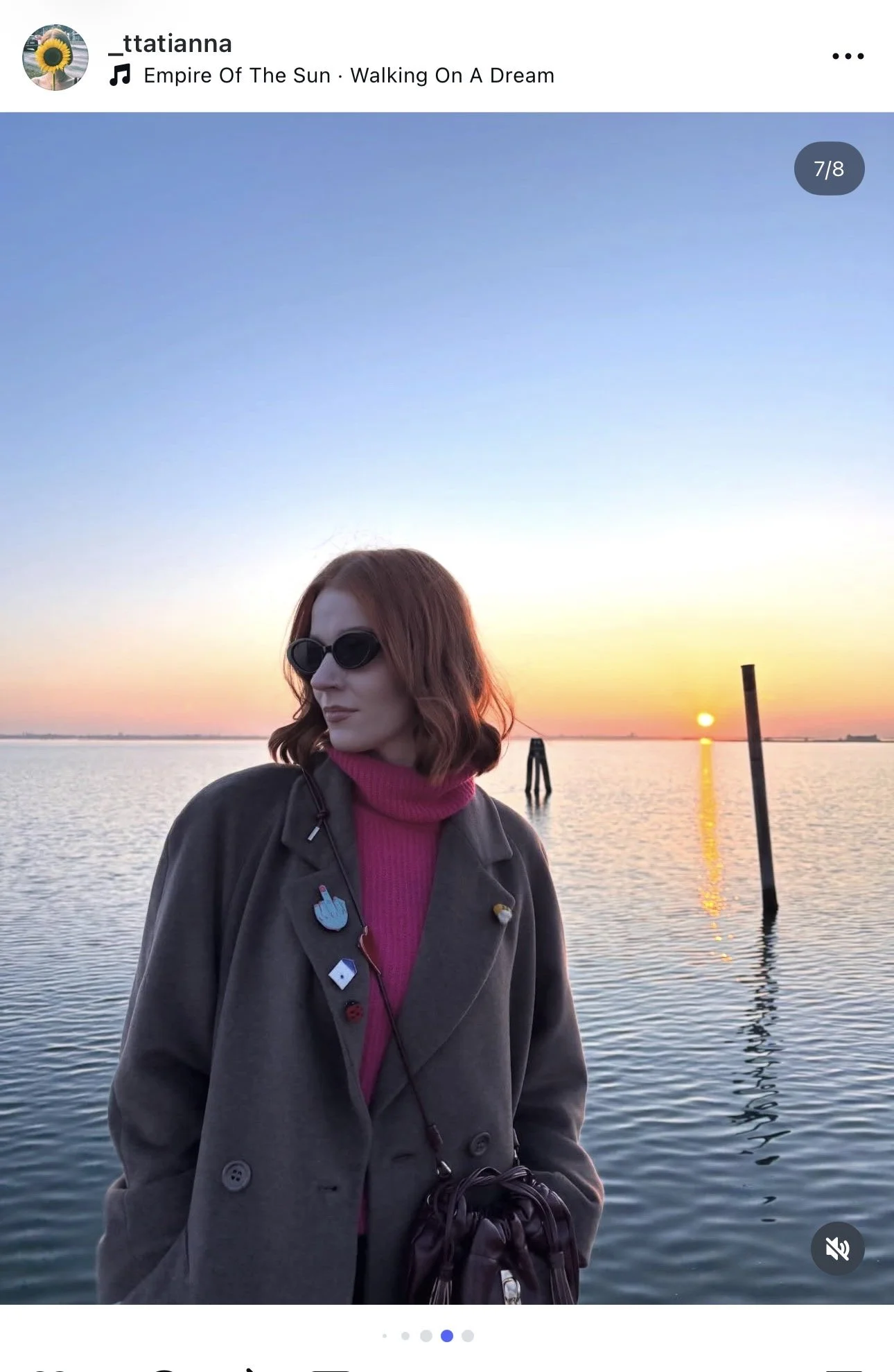 Woman with red hair wearing sunglasses and a gray coat with pins, standing by water during sunset, with a dock and the sun visible in the background.