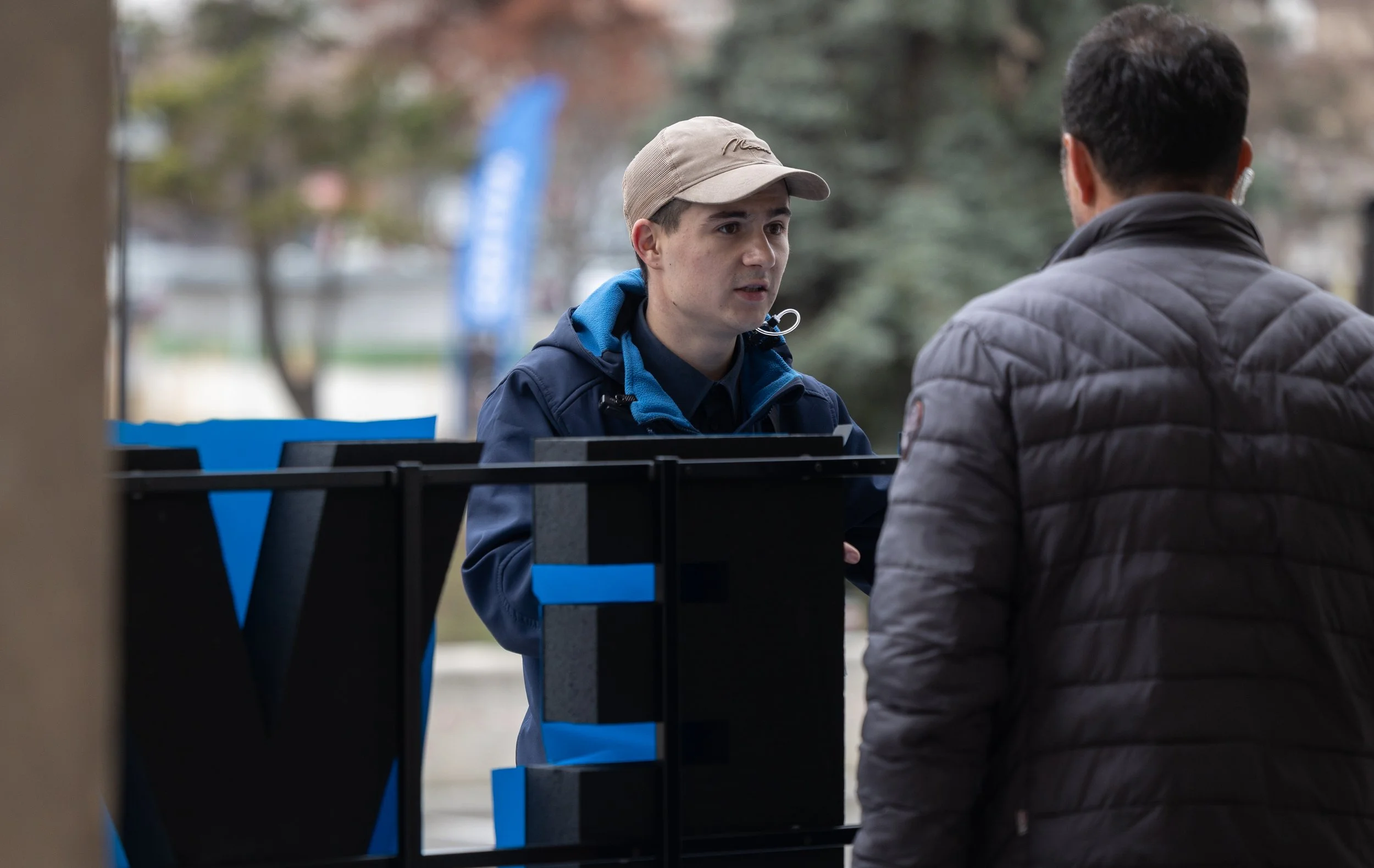 Two men are having a conversation outdoors, seen from behind and the front, with a background of trees and a park. One man is wearing a beige cap, a dark jacket with blue accents, and the other man has dark, slightly curly hair and is wearing a quilted gray jacket.