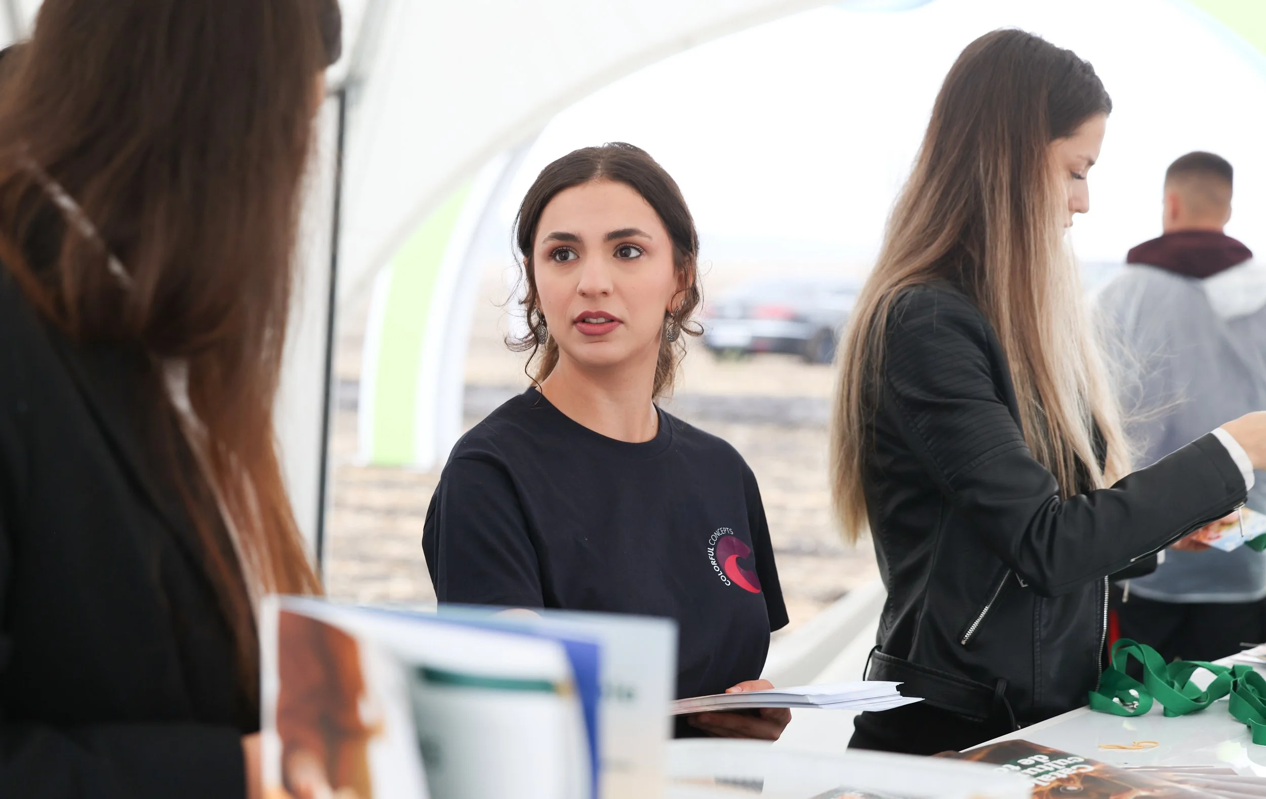 A woman with dark hair and earrings, wearing a black t-shirt with a logo, speaking to a group at an outdoor table under a canopy. Other people around her are engaged and blurred in the background.