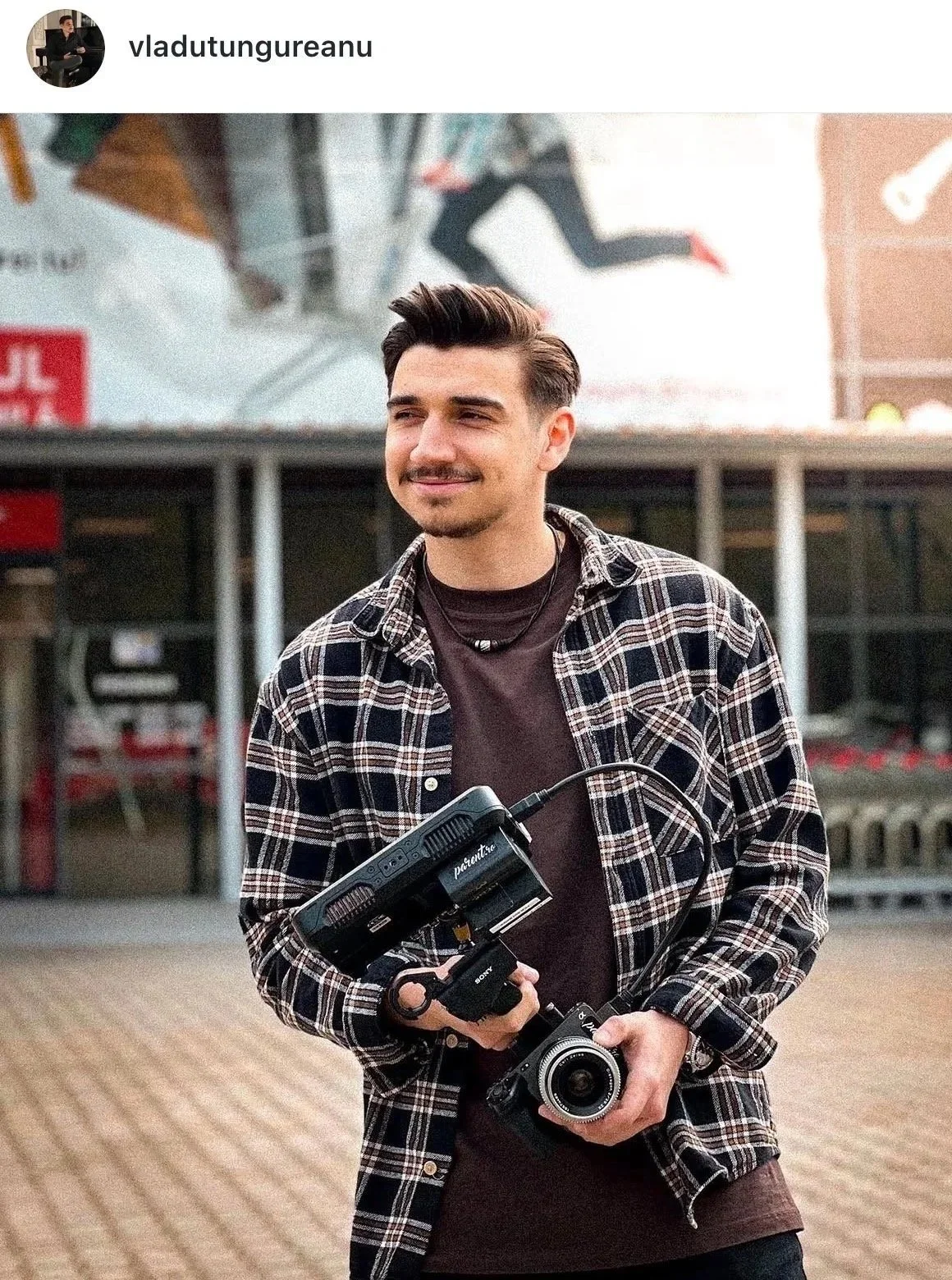A young man with dark hair and a goatee holds a camera with a stabilizer in his right hand, wearing a plaid shirt over a dark T-shirt, standing outdoors in an urban setting.