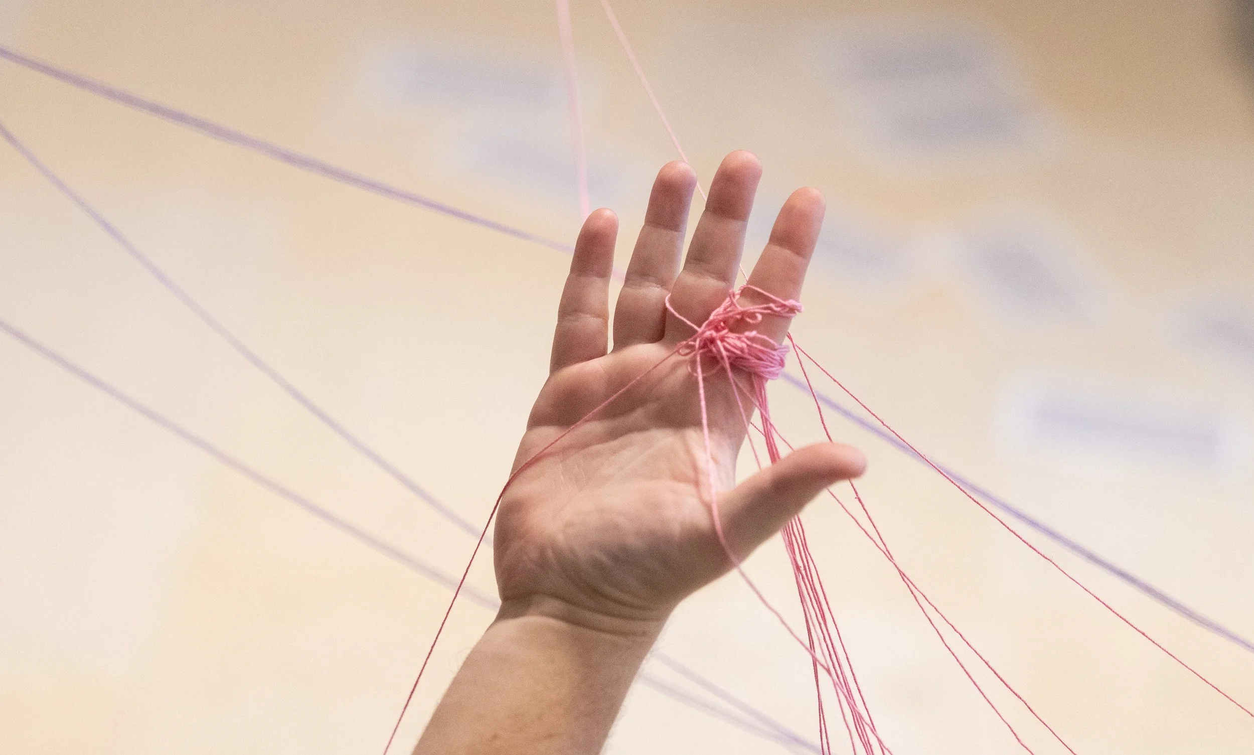 A person's hand with pink threads wrapped around fingers, with additional threads extending outward in a crisscross pattern against a pale background.