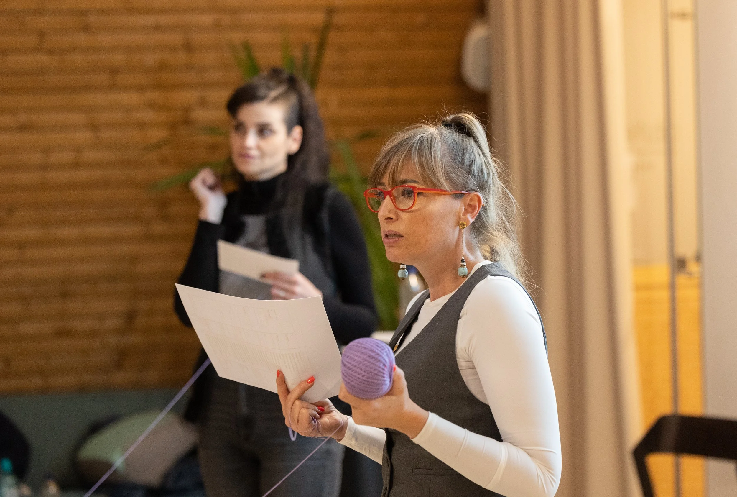 Two women are standing, holding papers, in a room with wooden wall paneling. One woman, in foreground, has blonde hair tied up, wears red glasses, a white shirt, and a gray vest with earrings. The other woman, in background, has dark hair styled in a partial mohawk, wears a black turtleneck, and is slightly blurred.