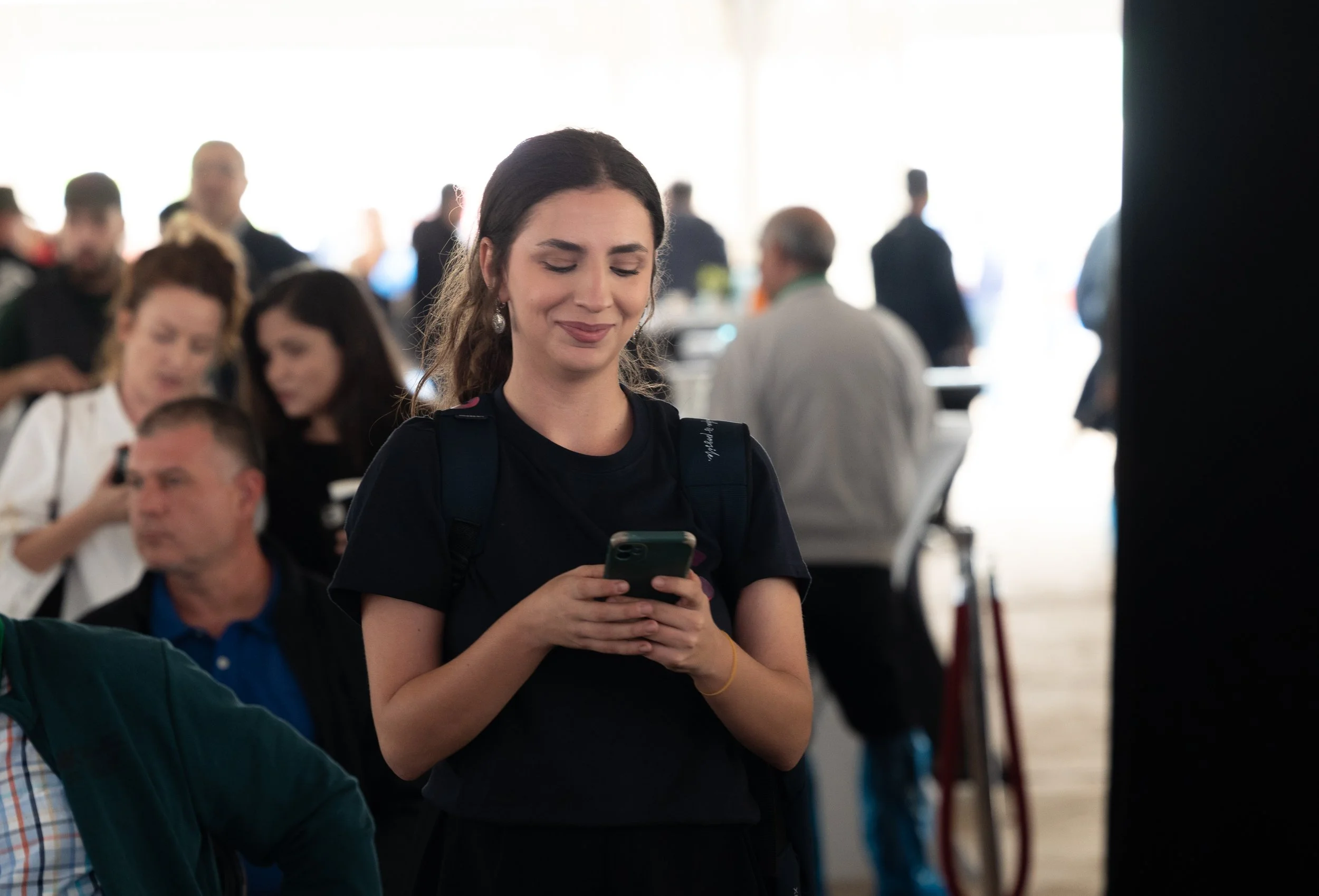 A woman standing and smiling while looking at her phone in a crowded indoor setting.