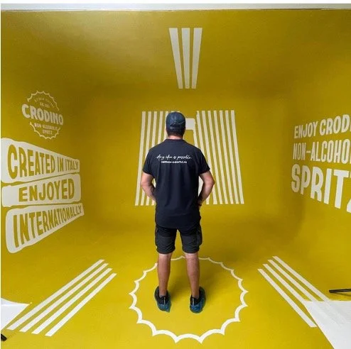 Man standing in yellow photo booth with white stripes and text promoting Crodino non-alcoholic spritz, designed with an optical illusion of platform and Vendome-style stripes.