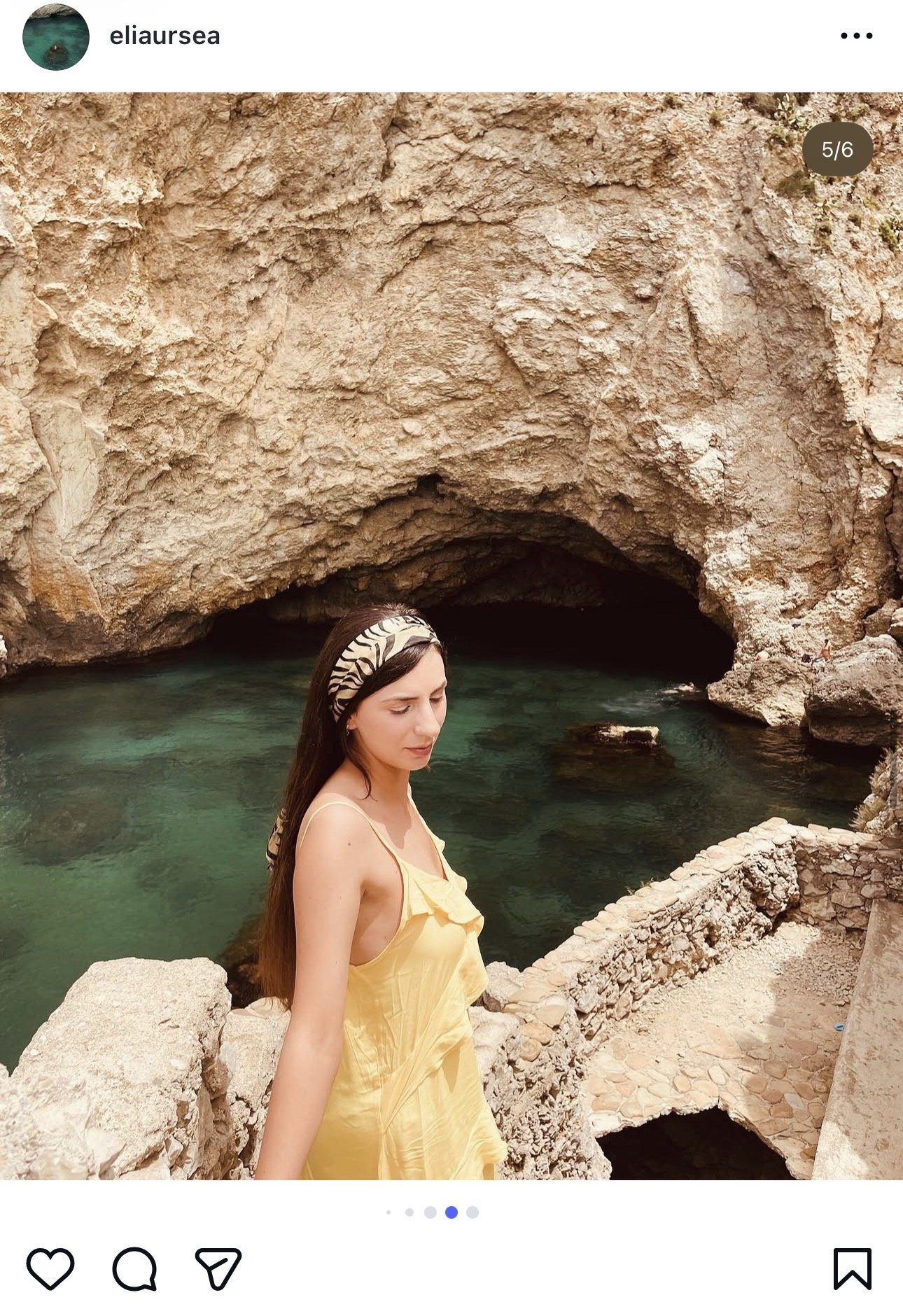A woman in a yellow dress standing near a rocky pool of water with a cave in the background.