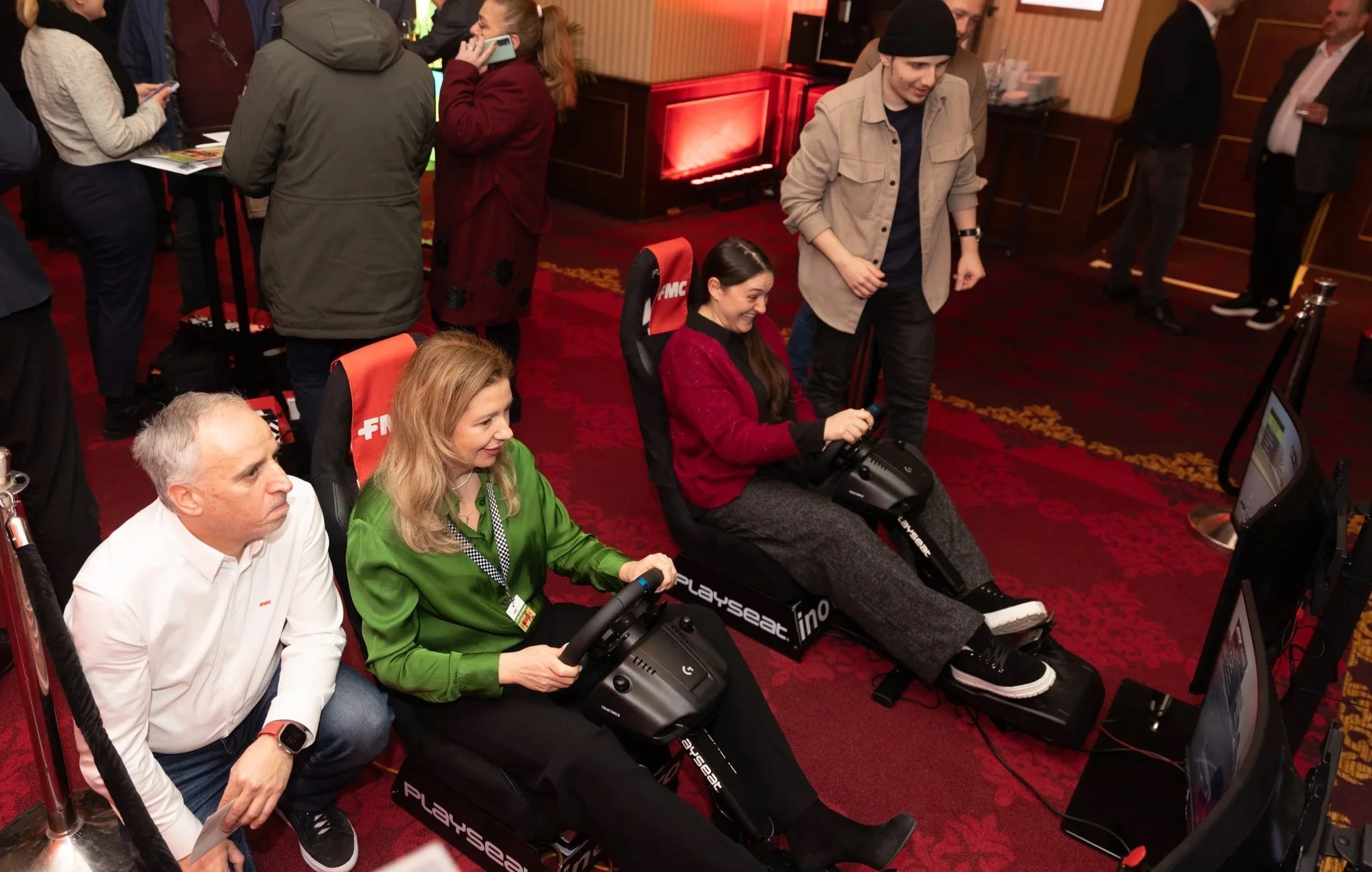 A group of people at an indoor event with some sitting in gaming simulation chairs holding controllers and others standing. The room has red carpet, patterned wallpaper, and a red piano in the background.