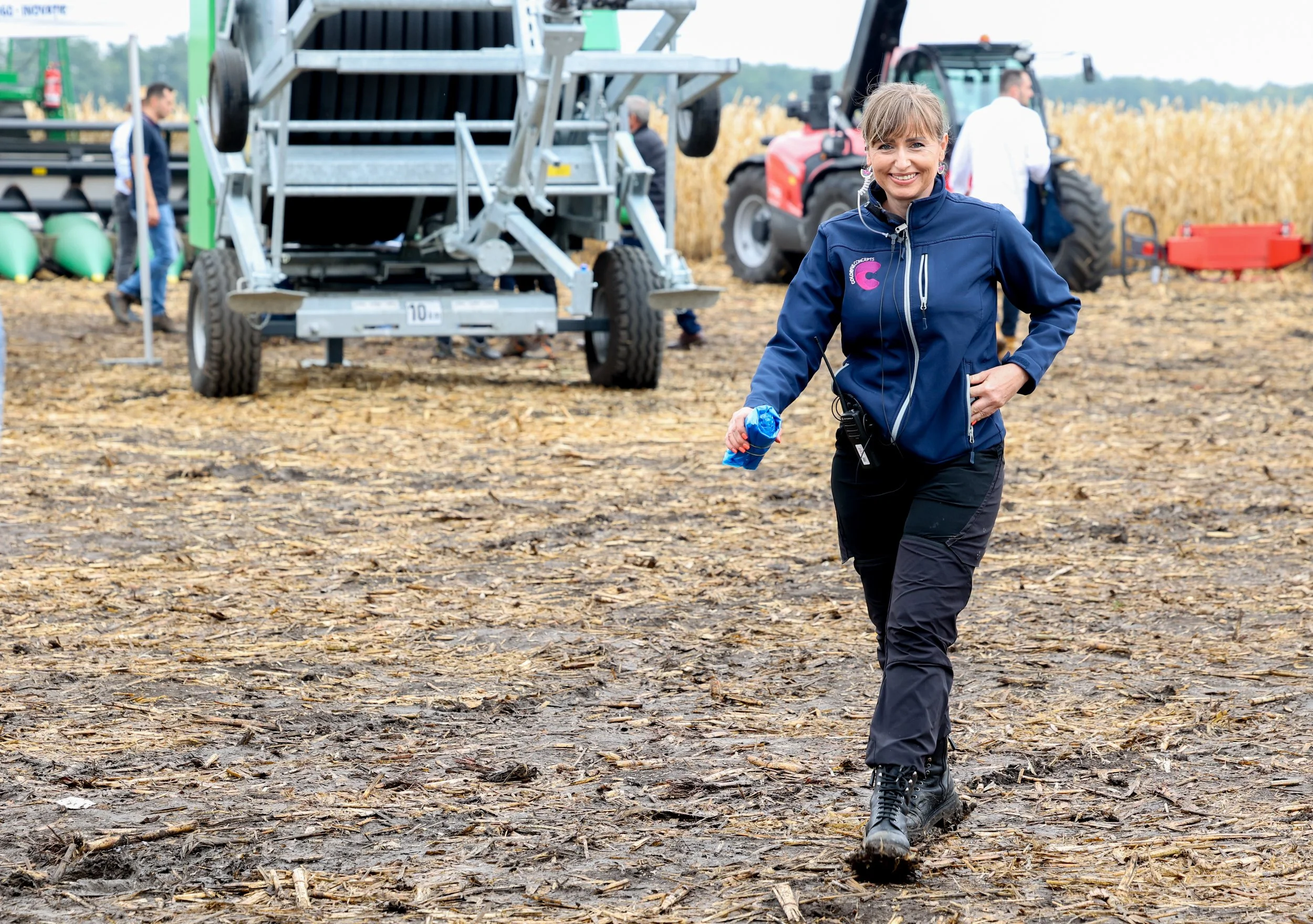 A woman dressed in outdoor attire, smiling and walking on a muddy farm field near farm equipment and a tractor, with a field of crops in the background.