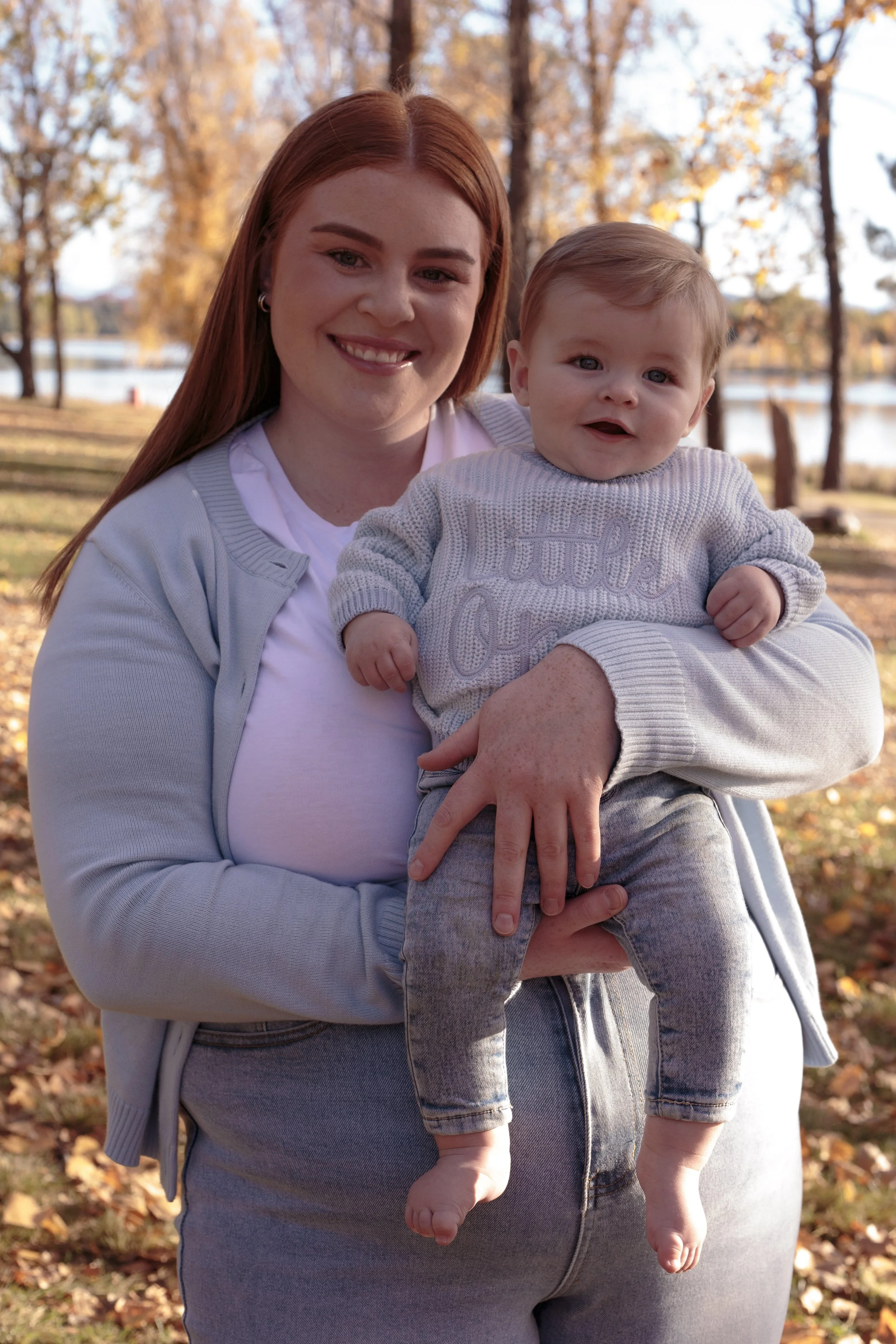 A woman with red hair holding a young child outdoors during fall, with trees and falling leaves in the background.