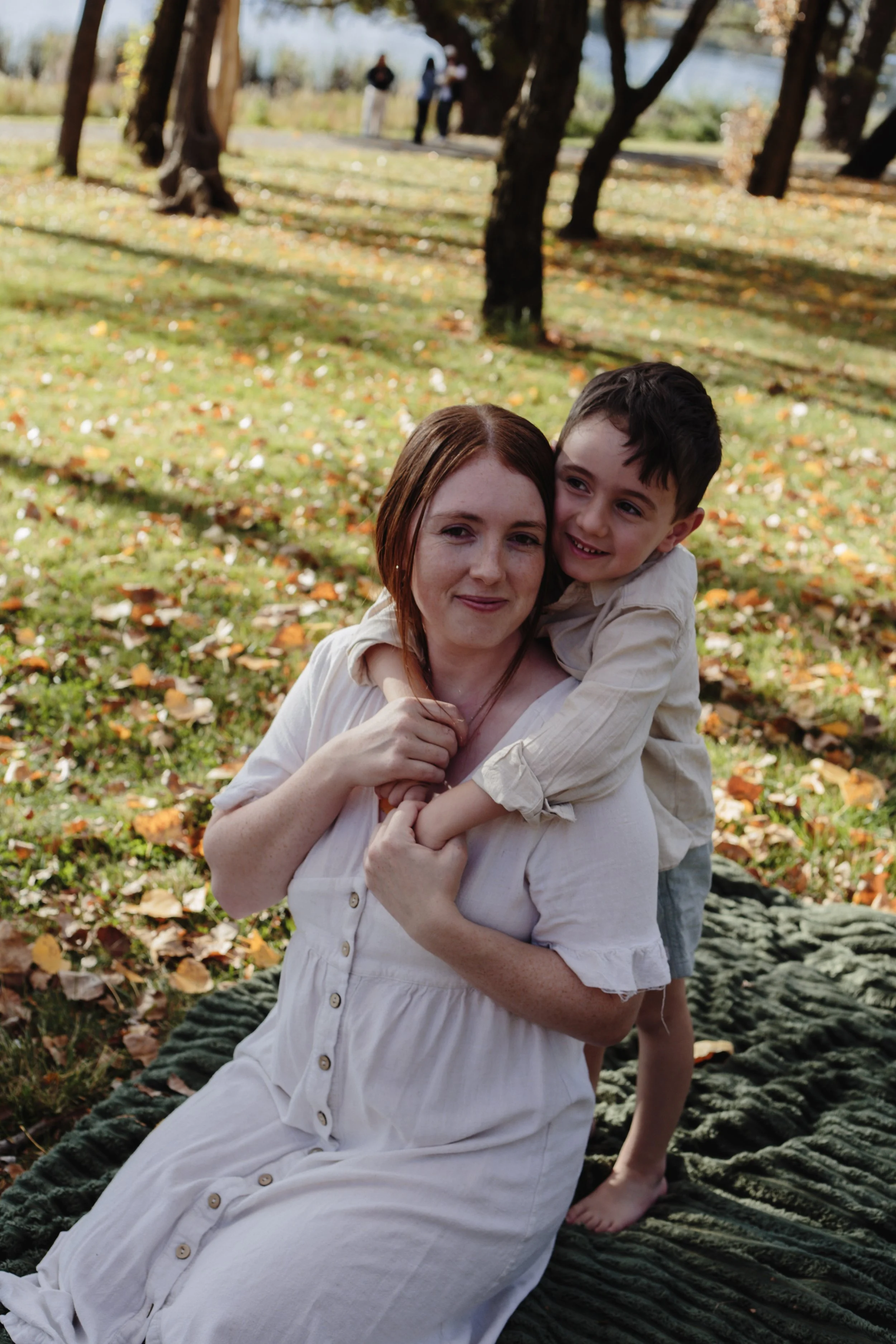 A woman and a young boy outdoors in a park with fallen autumn leaves. The woman is sitting on a large blanket, and the boy is hugging her from behind, smiling.