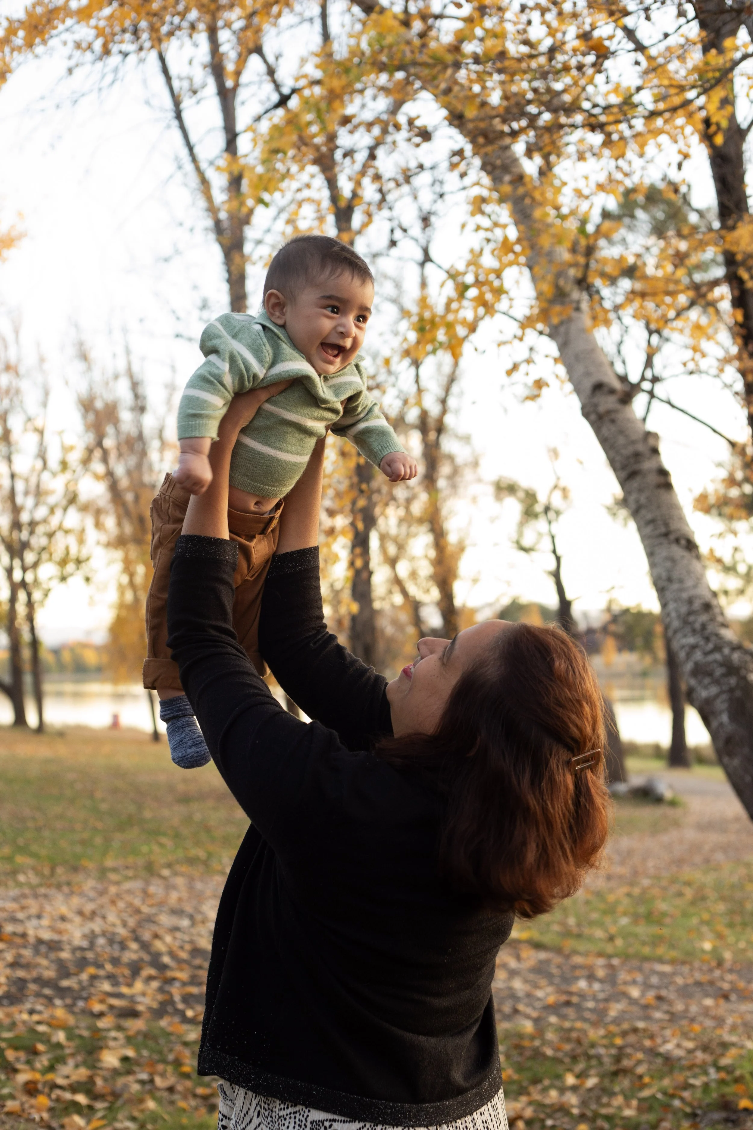 A woman lifting a happy toddler boy with a background of autumn trees with yellow and orange leaves.