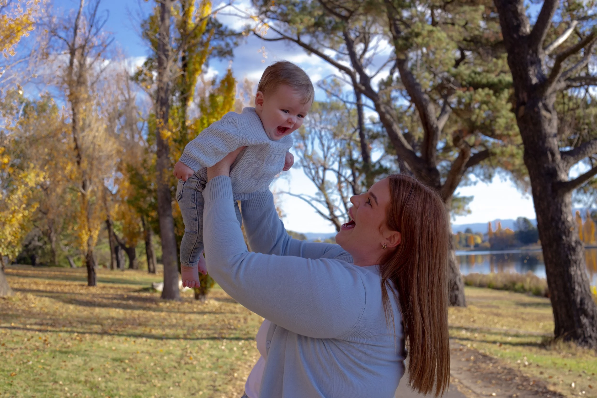 A woman lifting a young child in the air outdoors in a park during autumn. The woman is smiling, and the child appears excited. The background features trees with colorful fall leaves and a blue sky with some clouds.