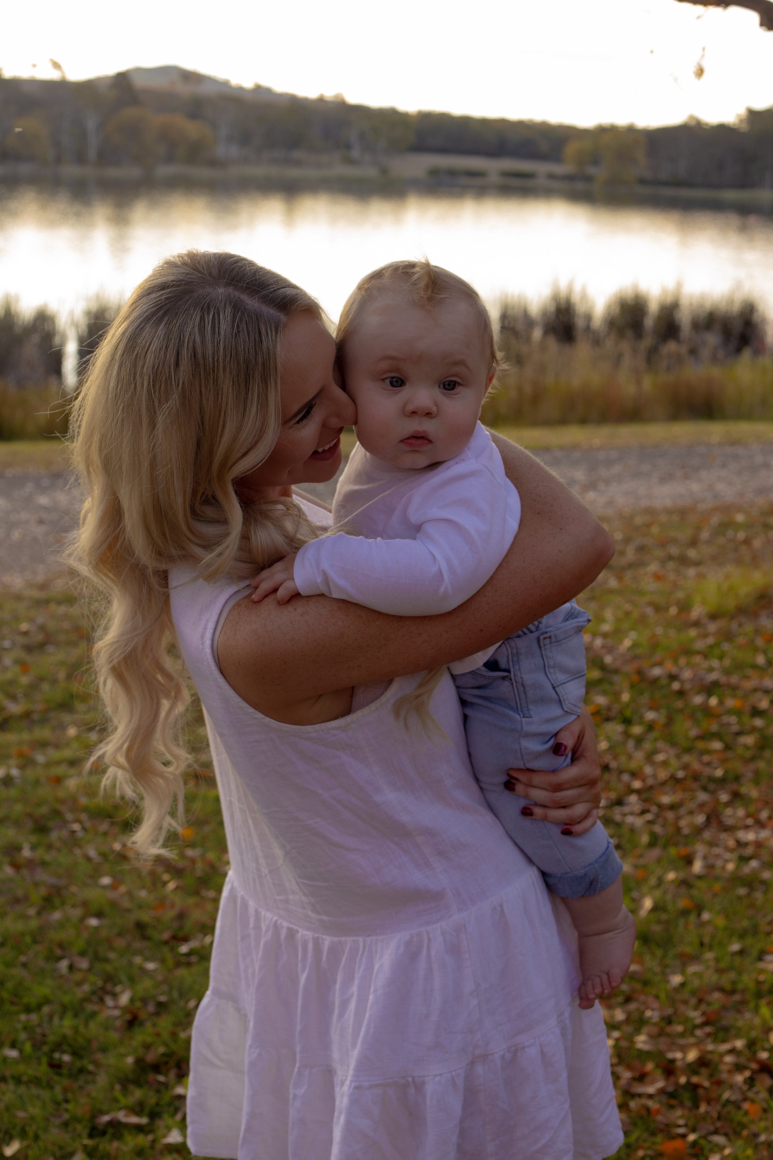 A woman with long blonde hair holding a young boy outdoors near a lake, with autumn leaves on the ground and trees in the background.