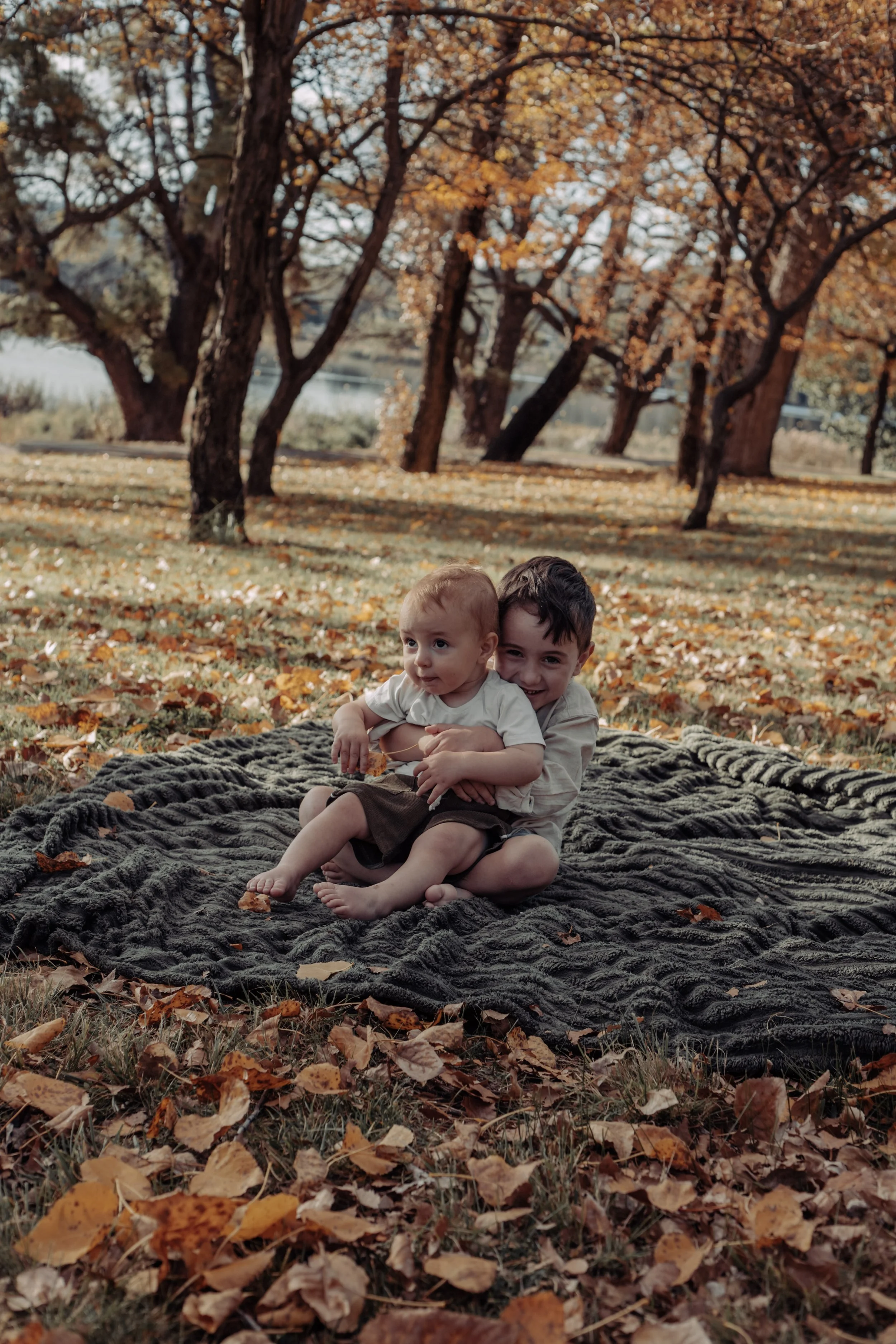 Two children sitting on a blanket on the ground surrounded by fallen autumn leaves, with trees in the background.