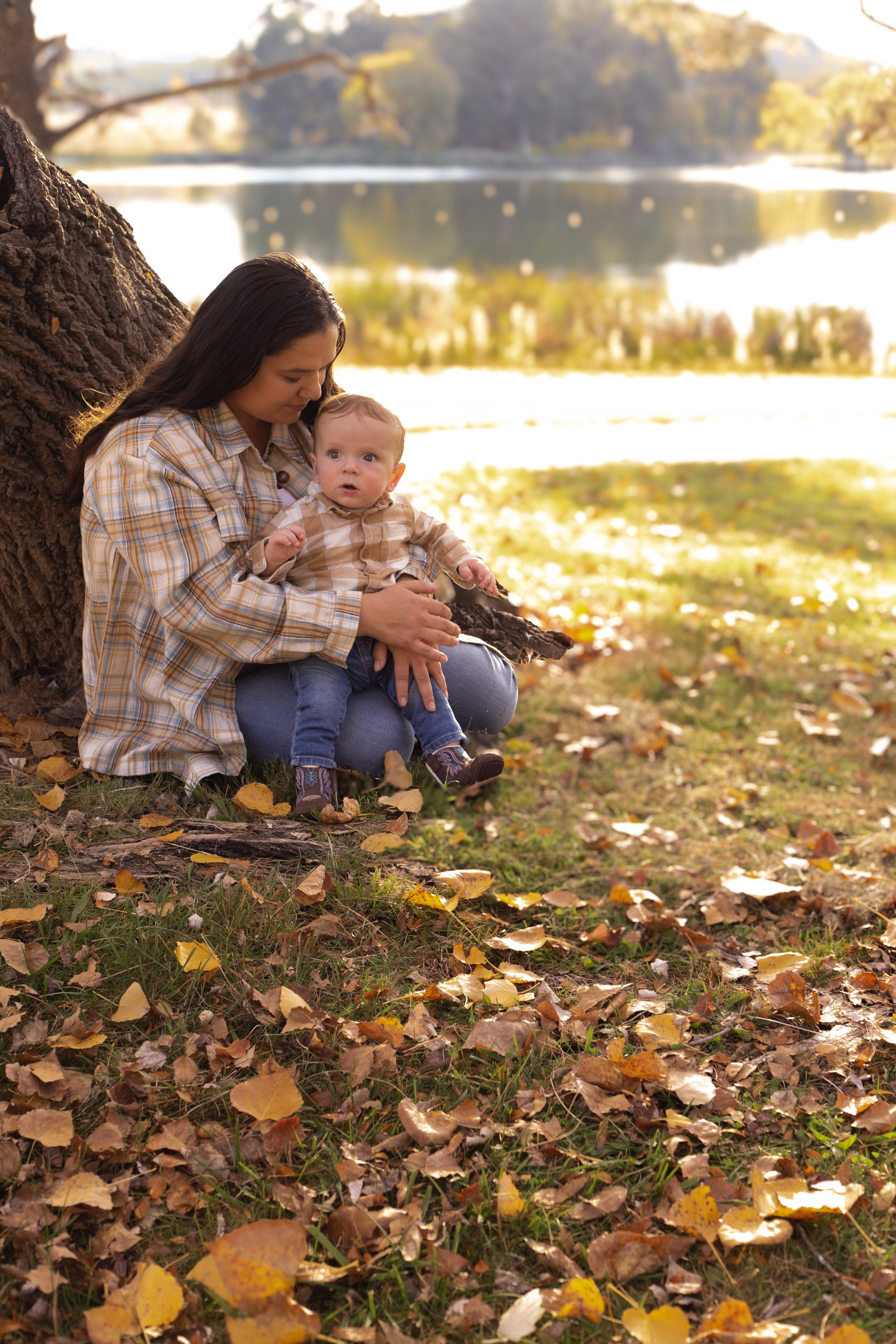 A woman and a young boy sitting under a large tree by a lake during autumn, with fallen leaves on the ground and a blurred landscape of trees and water in the background.