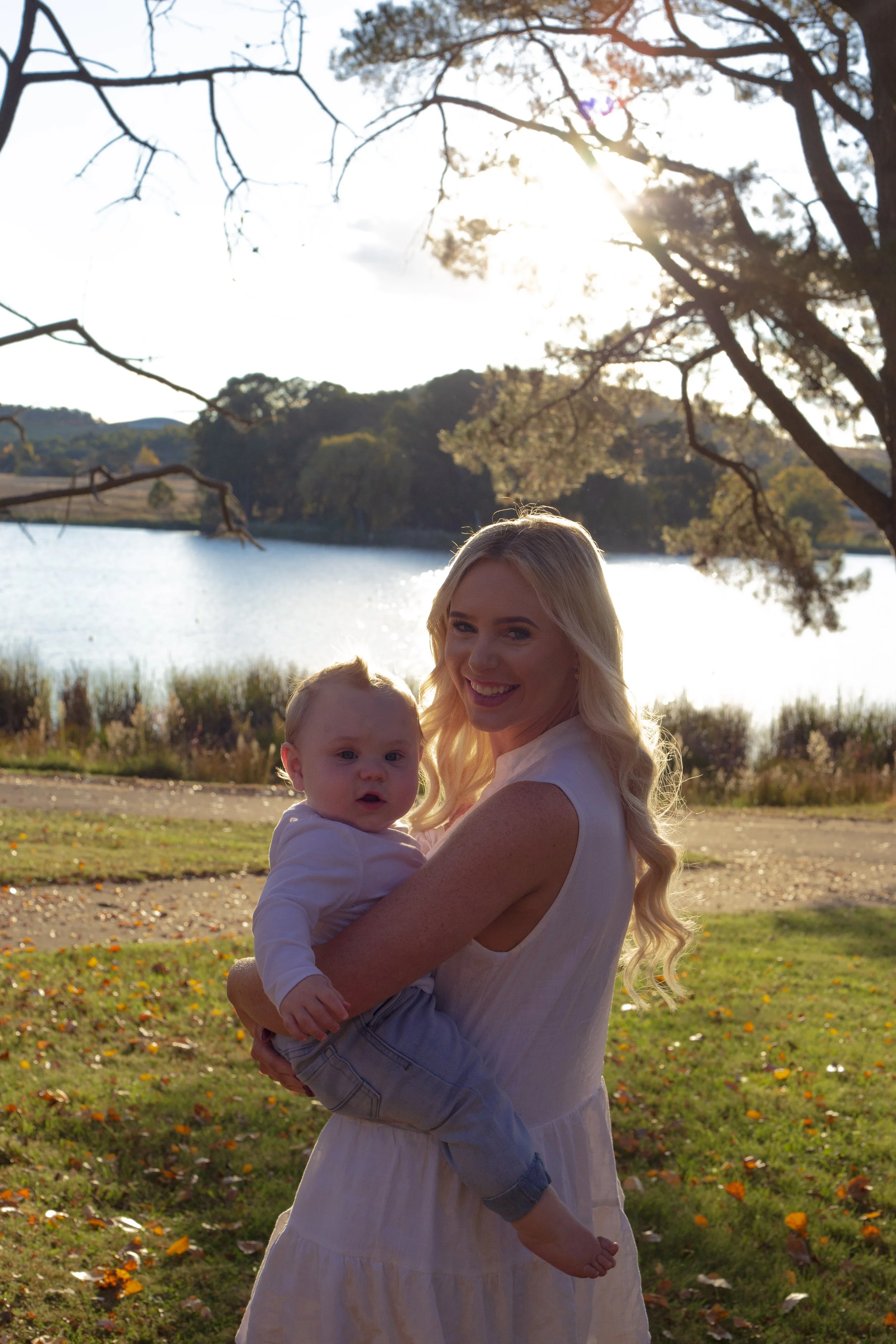 A woman with long blonde hair holding a baby outdoors near a lake with trees and hills in the background, during sunset.