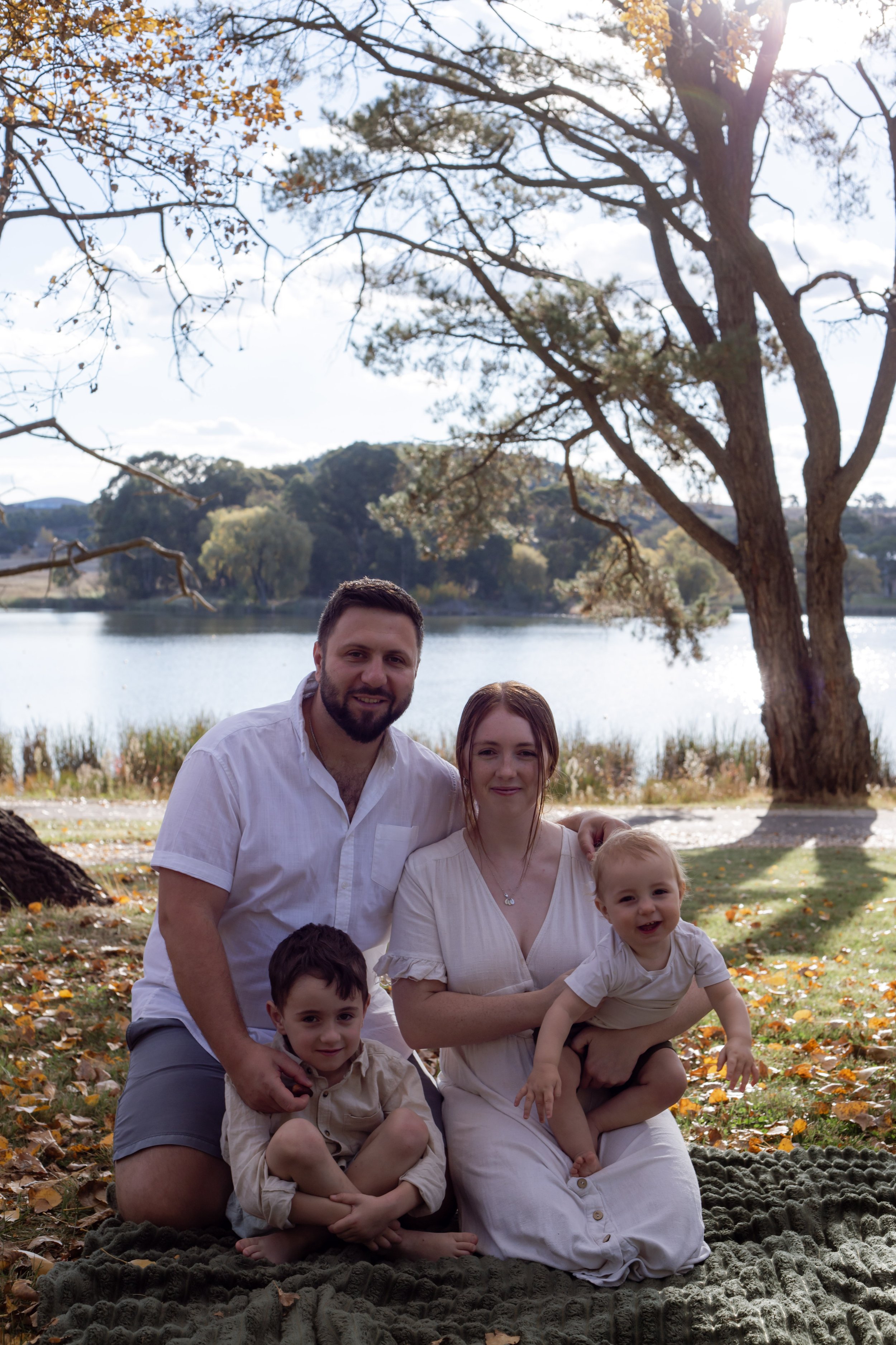 A family of five posing outdoors in a park beside a lake with trees showing autumn colors. The adults and children are smiling, and the children are sitting on a blanket on the ground.
