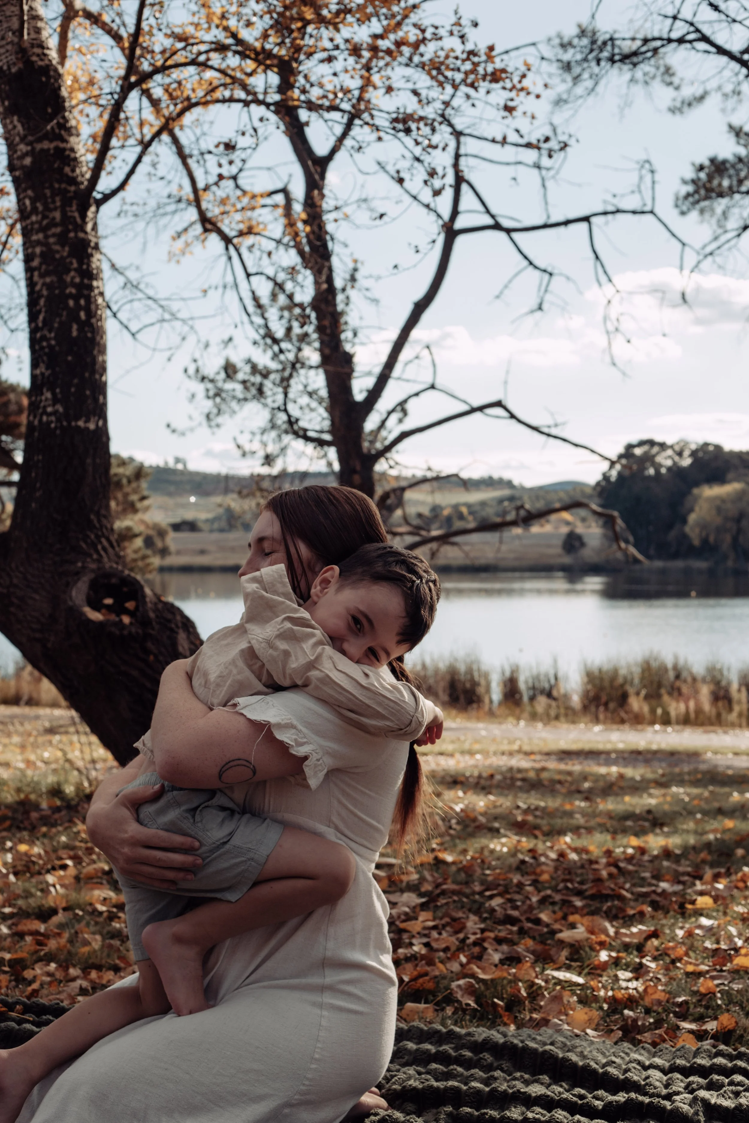 A woman and a young boy hugging outdoors near a lake, with trees and fallen autumn leaves in the background.