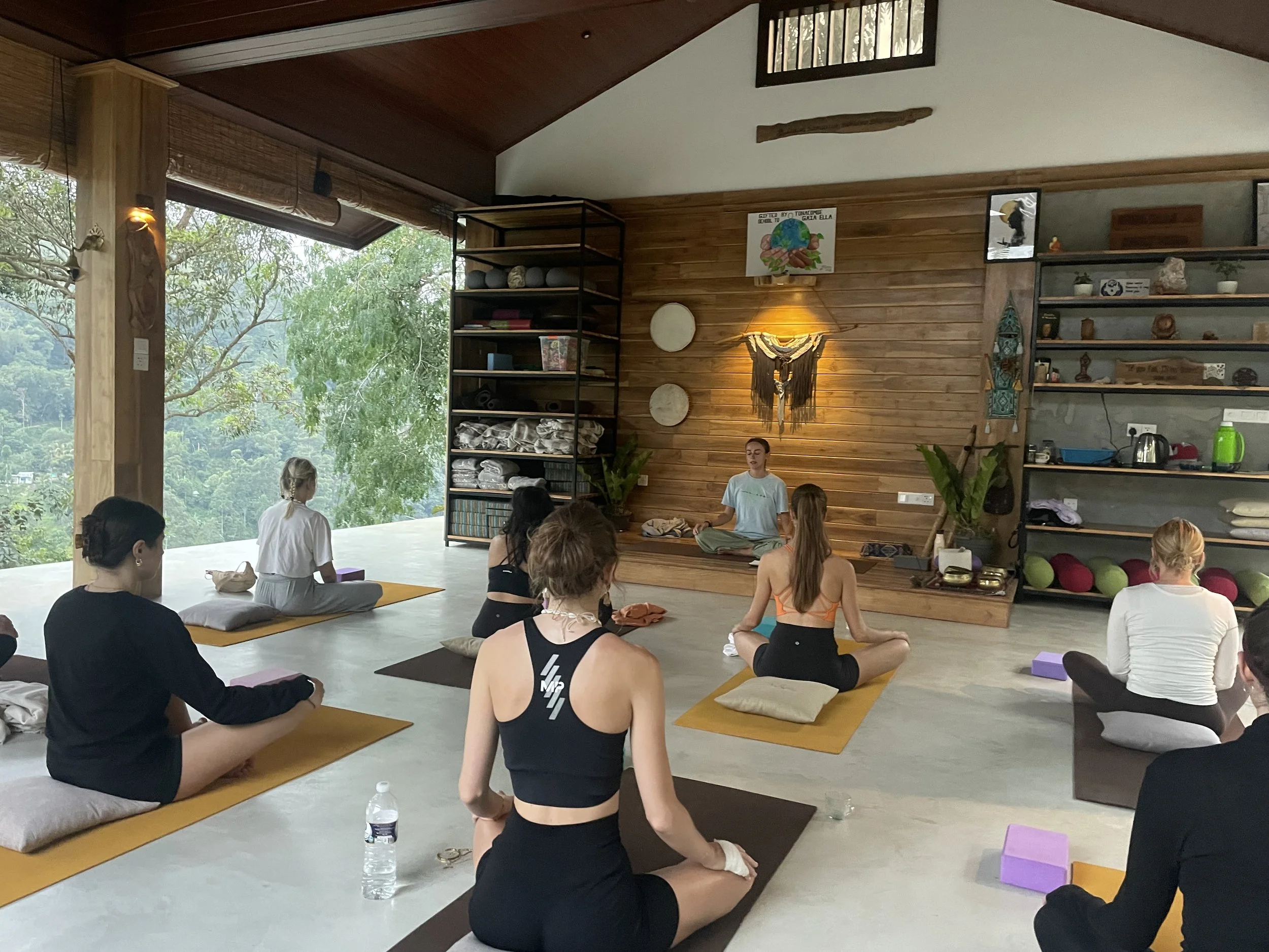 A group of people participating in a yoga or meditation class in a room with large windows, wooden walls, and shelves with various items, sitting on yoga mats in a cross-legged position.