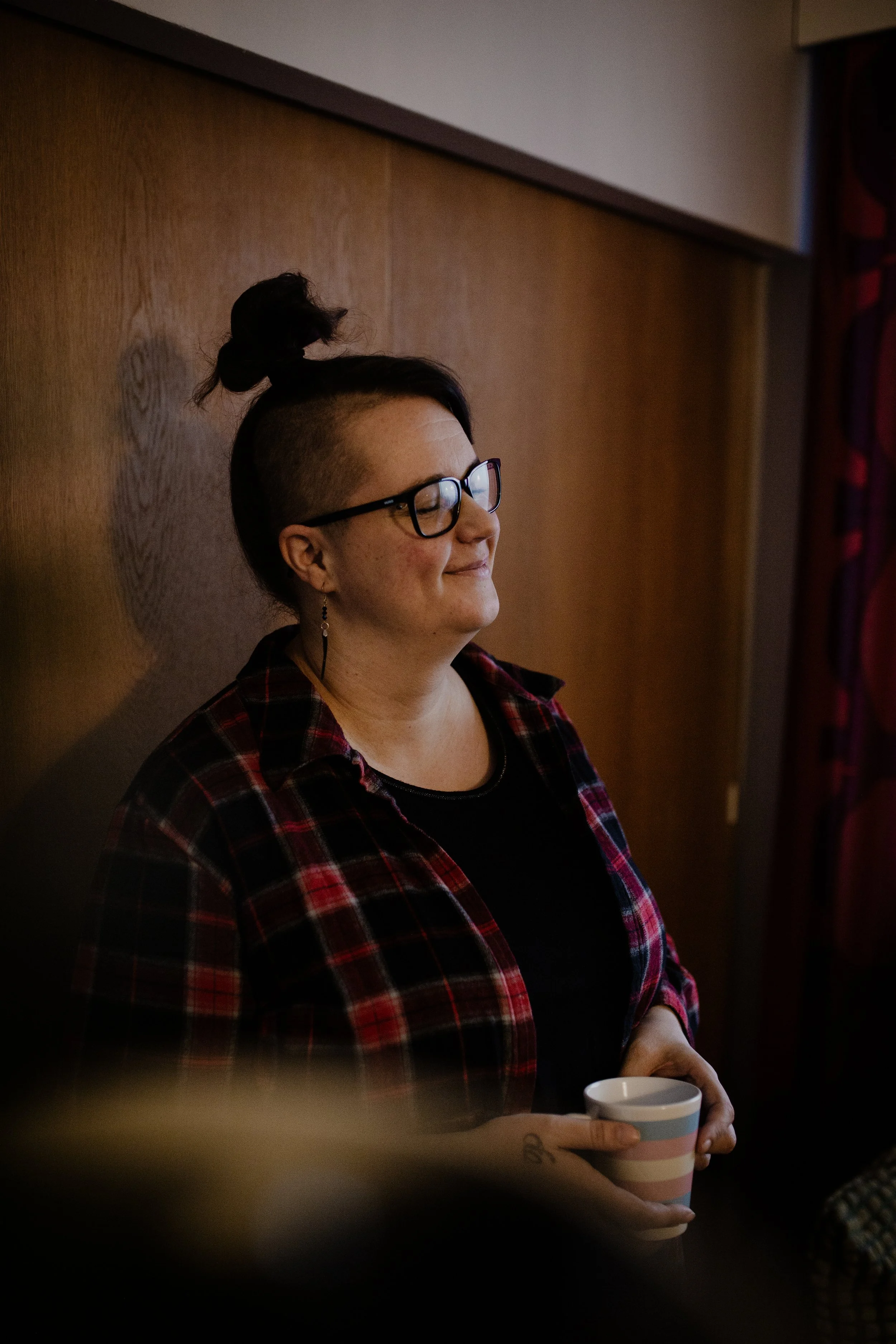Ihminen pitää kädessään kahvimukia ja seisoo taustalla puupaneeliseinässä, silmät suljettuina ja hymyillen.