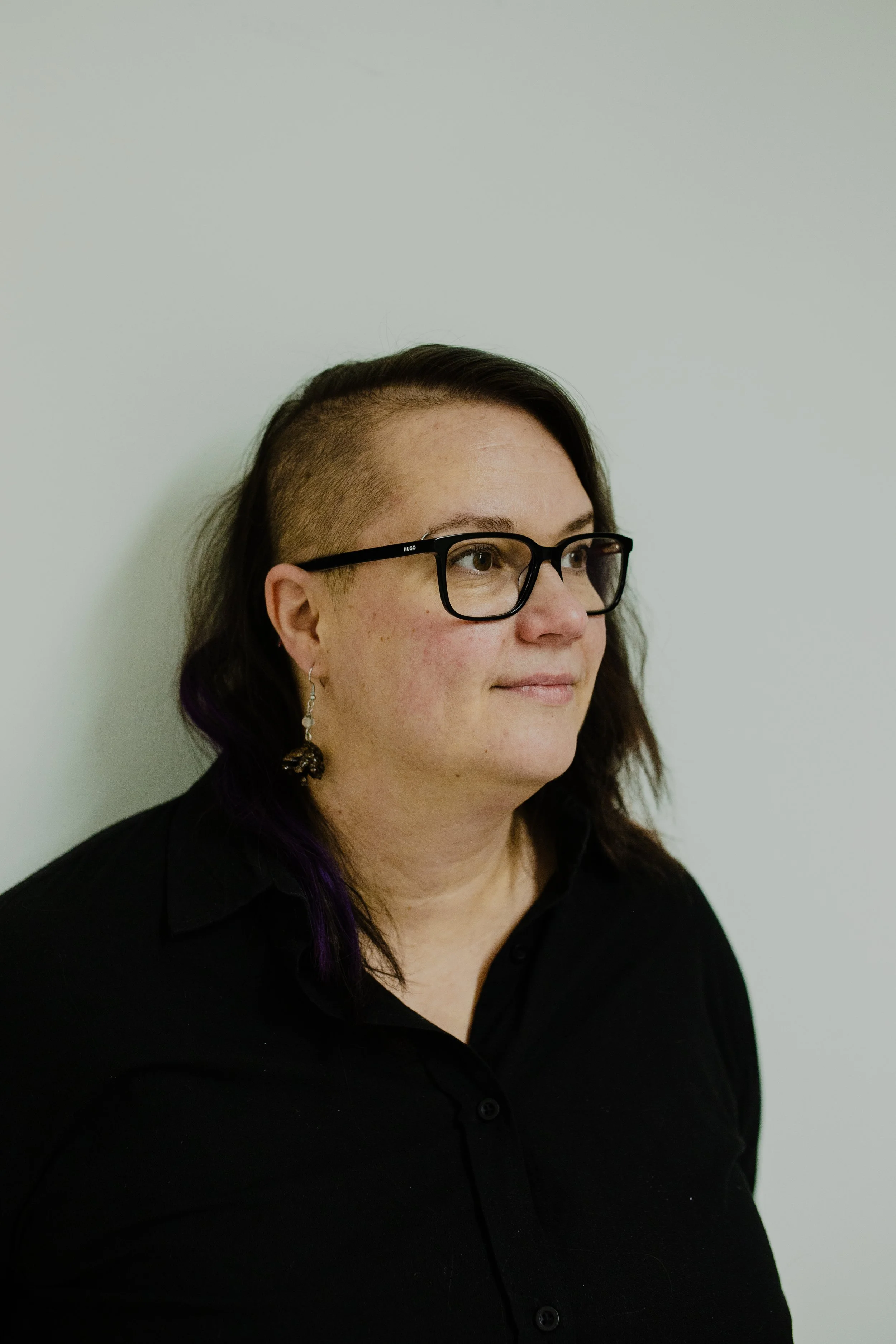 Nainen, jolla on lyhyt hiusleikkaus toiselta puolelta ja pitkä toiselta, käyttää silmälaseja ja mustaa paitaa, taustalla vaaleanharmaa seinä.
