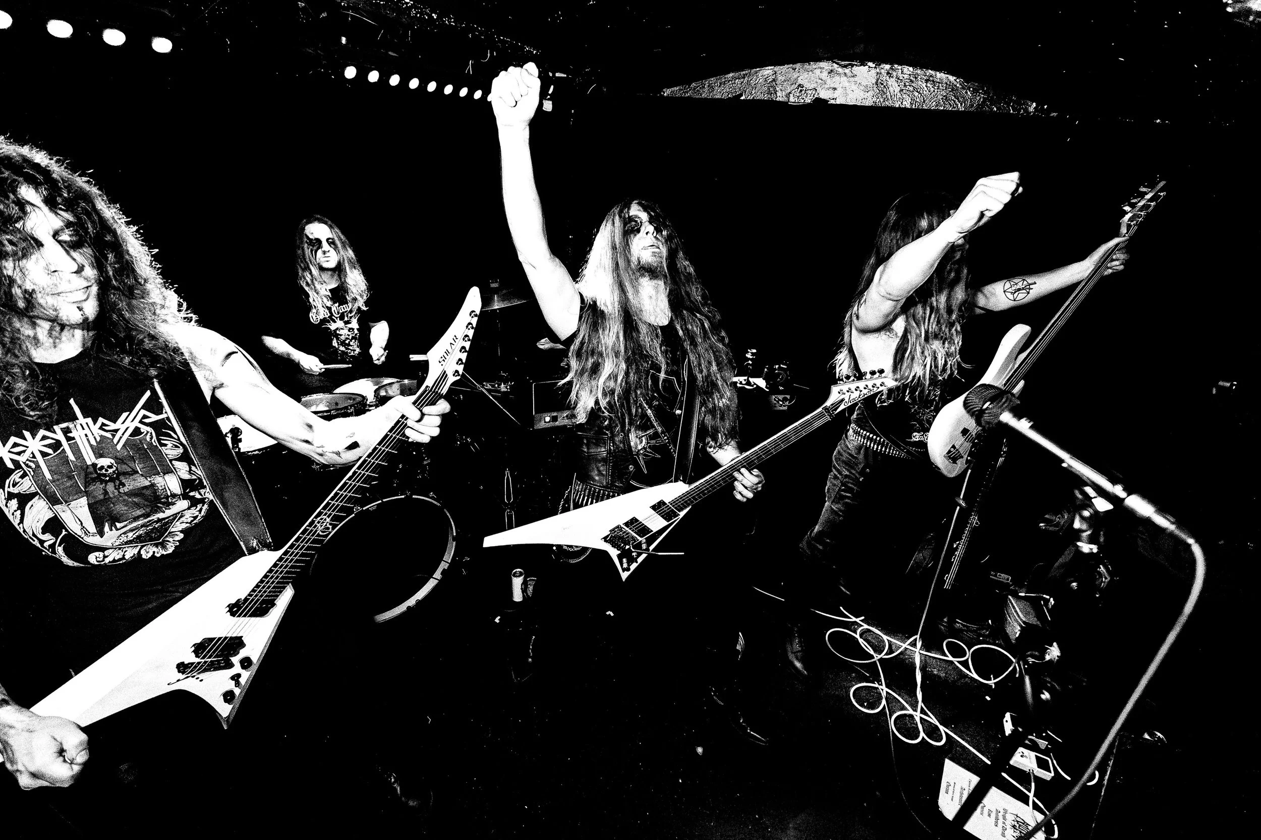 A black and white photo of a band performing live, featuring three musicians with long hair playing guitars and a drummer in the background. One musician is raising a fist.