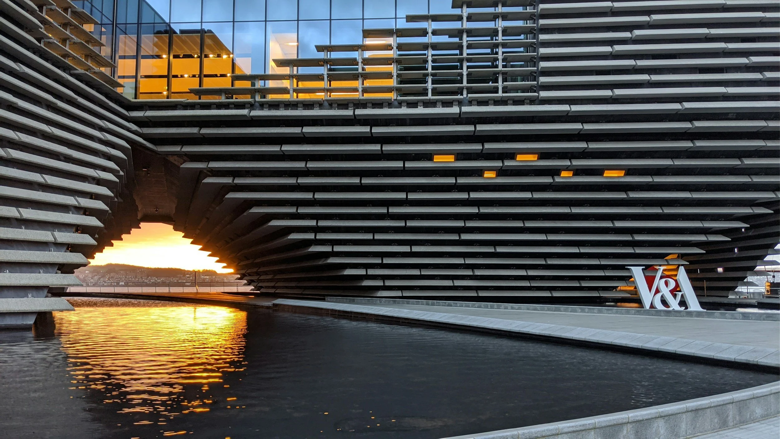Modern building with horizontal slats and glass windows, reflecting sunset water, featuring an arch-shaped gap at the bottom and a 'V&A' sign.  Heather Ogilvie and The Ogilvie Business School in Dundee, Scotland.