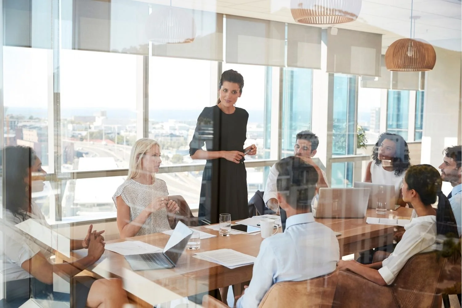 Business meeting in a modern conference room with large windows and city view, featuring a woman standing and presenting to a seated diverse group of professionals. Heather Ogilvie and The Ogilvie Business School in Dundee, Scotland.