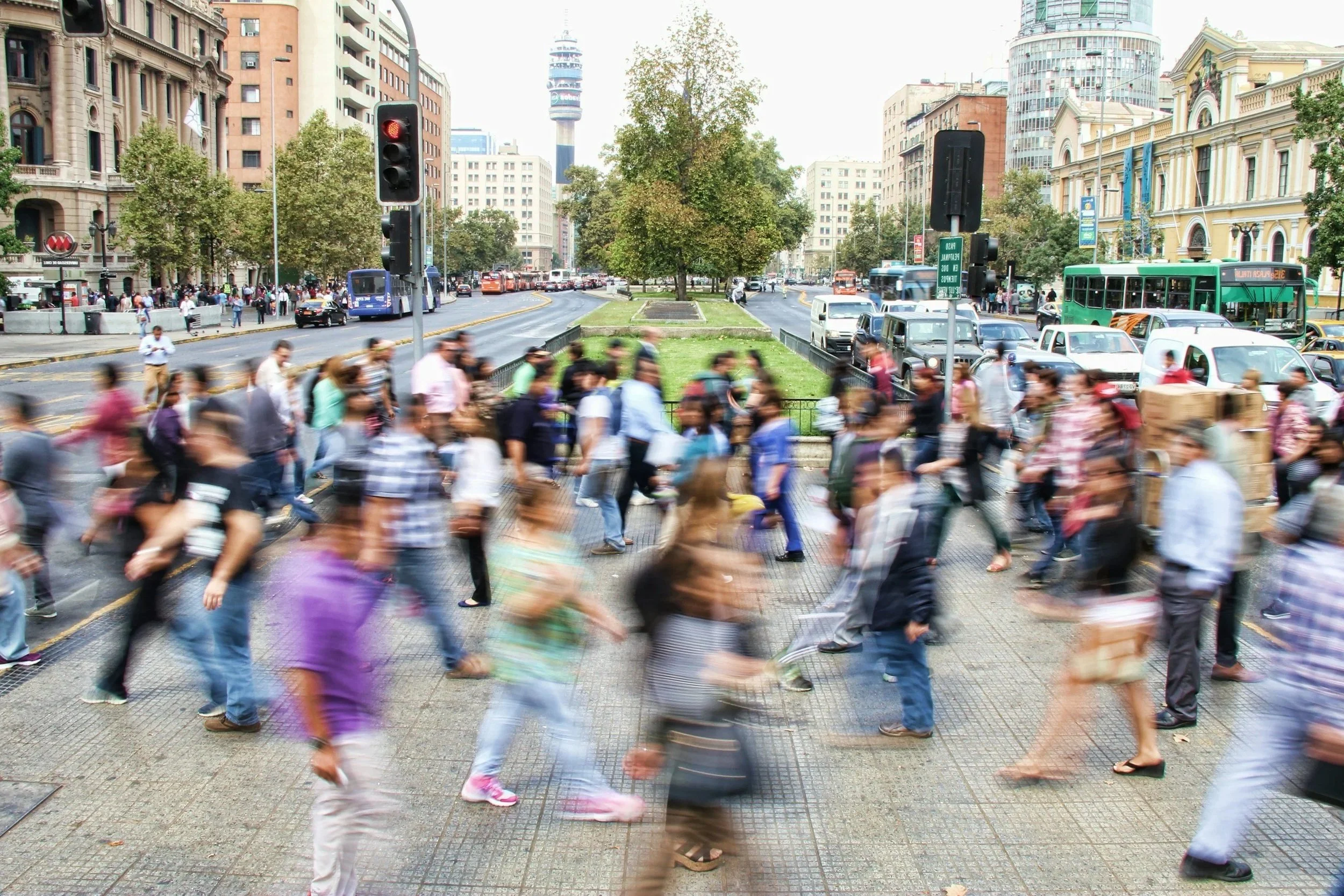 A busy city street with a large crowd of pedestrians crossing at a crosswalk, buildings, trees, and traffic including buses, cars, and a green streetcar, in an urban setting.  Heather Ogilvie and The Ogilvie Business School in Dundee, Scotland.