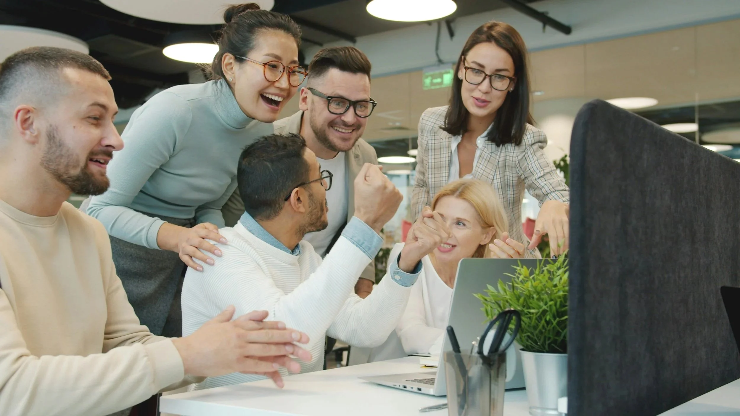 Group of six diverse colleagues gathered around computer monitor, celebrating and cheering in an office.  Heather Ogilvie and The Ogilvie Business School in Dundee, Scotland.