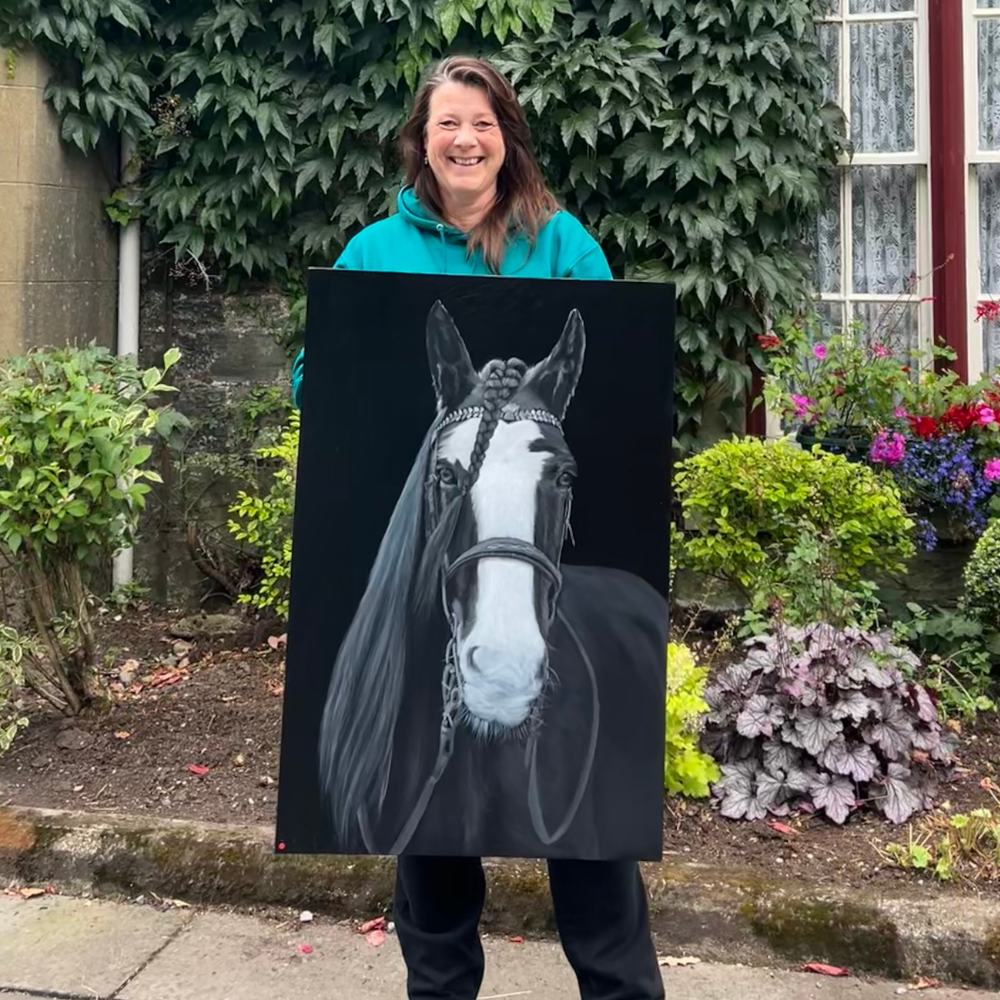 A woman smiling and standing outdoors in front of a garden with colorful flowers and green foliage, holding a large painting of a black and white horse.  Heather Ogilvie and The Ogilvie Business School in Dundee, Scotland.