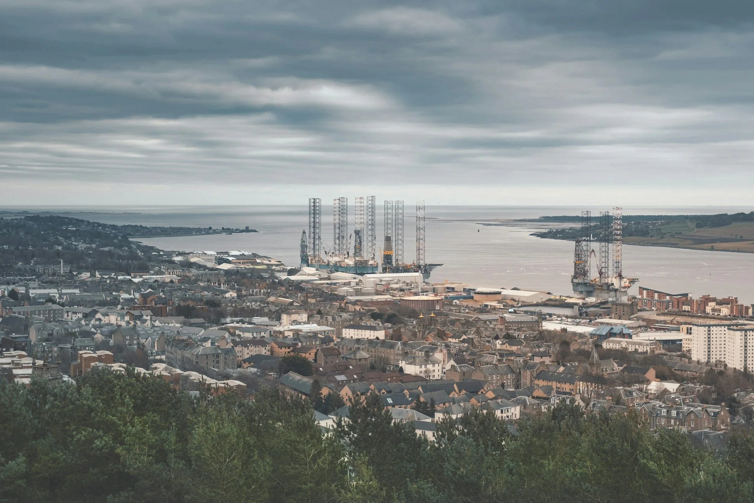 A cityscape view with a body of water in the background under a cloudy sky. Foreground includes residential houses and trees, with industrial structures and cranes near the water.  Heather Ogilvie and The Ogilvie Business School in Dundee, Scotland.