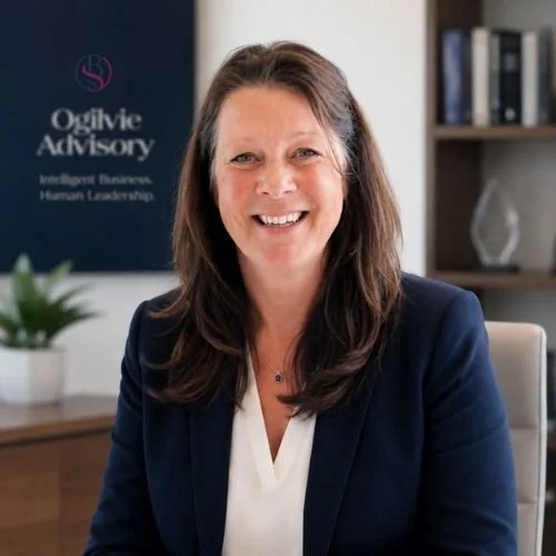 Professional woman with long brown hair smiling, sitting in an office with a bookshelf and a plant in the background. Heather Ogilvie and The Ogilvie Business School in Dundee, Scotland.