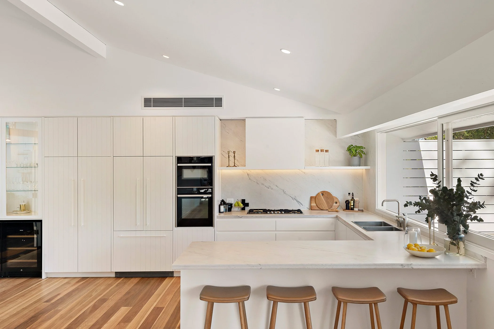 Modern white kitchen with a marble countertop, built-in oven, gas stove, and large window with a view of greenery. Decor includes a vase with greenery, a bowl of lemons, and kitchen utensils.