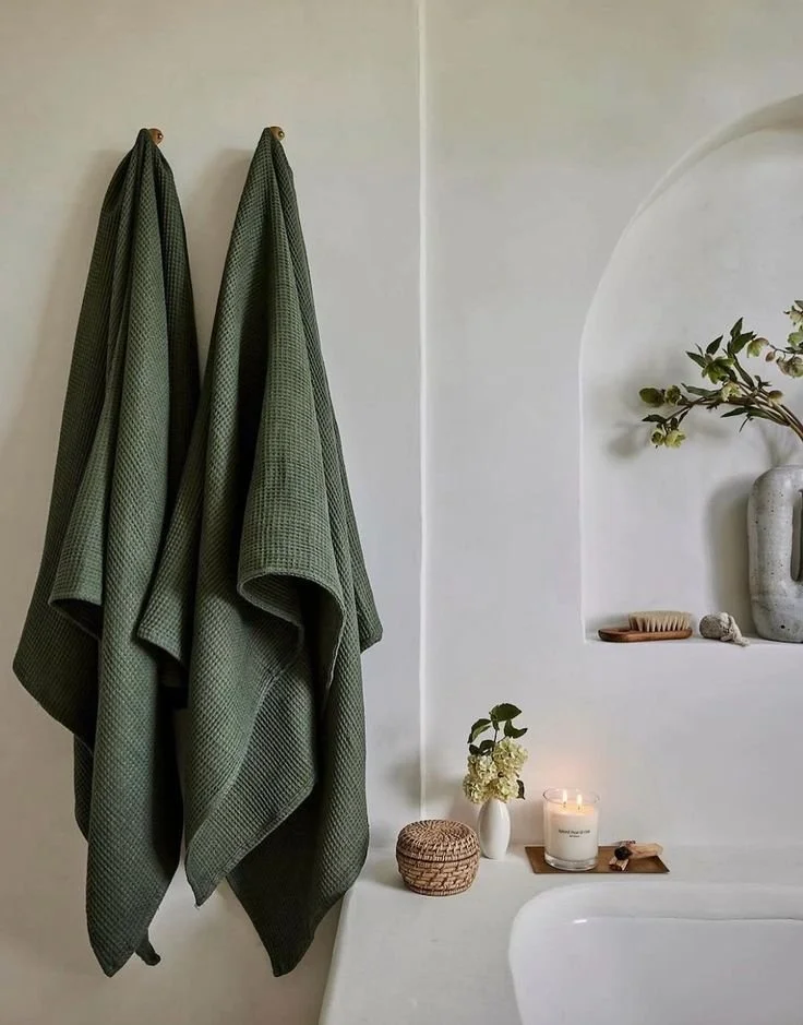 Two green towels hanging on hooks on a white wall with decor including candles, a vase with flowers, and small decorative items.