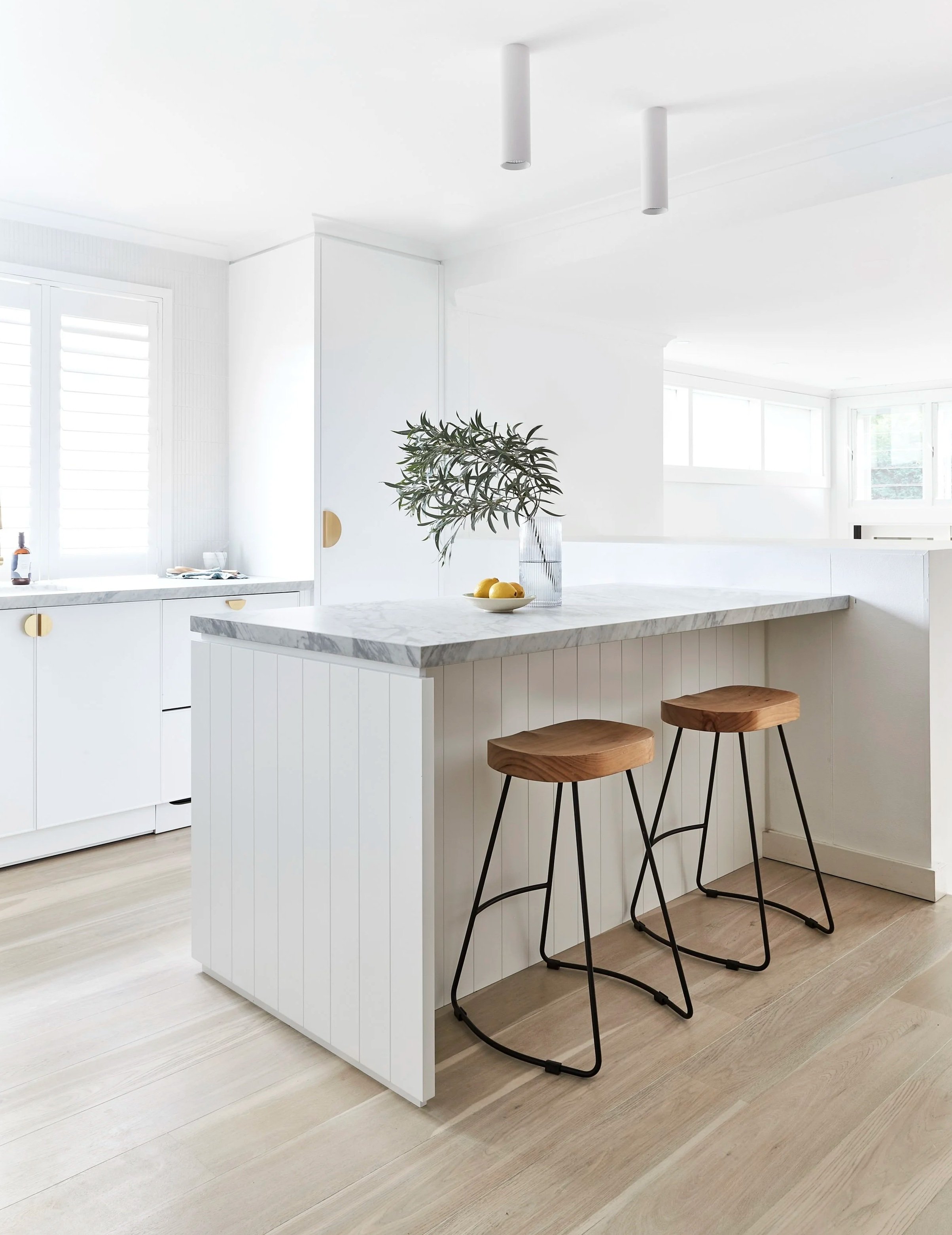 Modern white kitchen with a marble island, two wooden and black metal bar stools, a bowl of lemons, a glass vase with green foliage, and large windows letting in natural light.