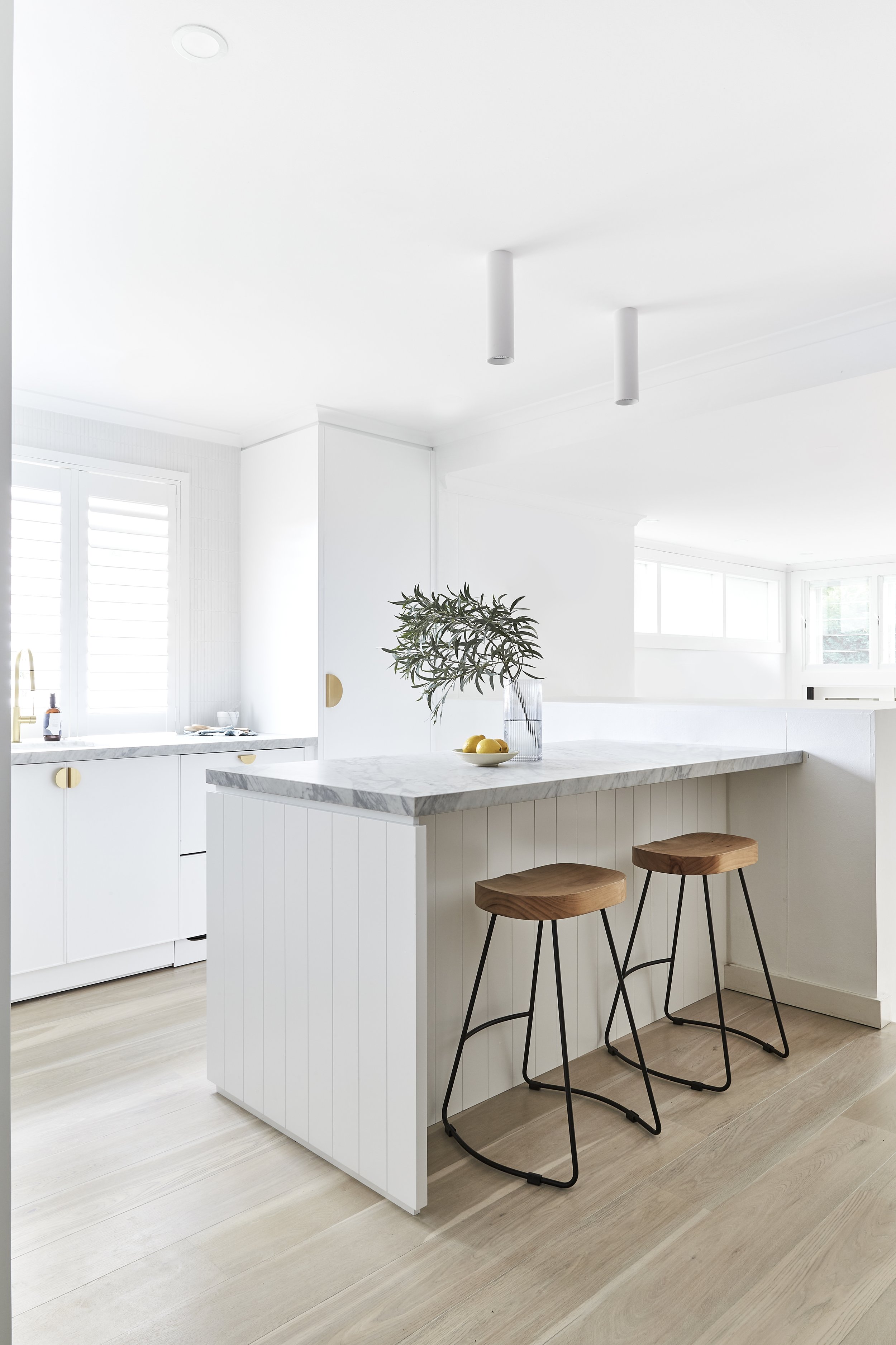 Minimalist white kitchen with a marble-topped island, three wooden bar stools with black metal legs, potted plant, and large windows with white shutters.