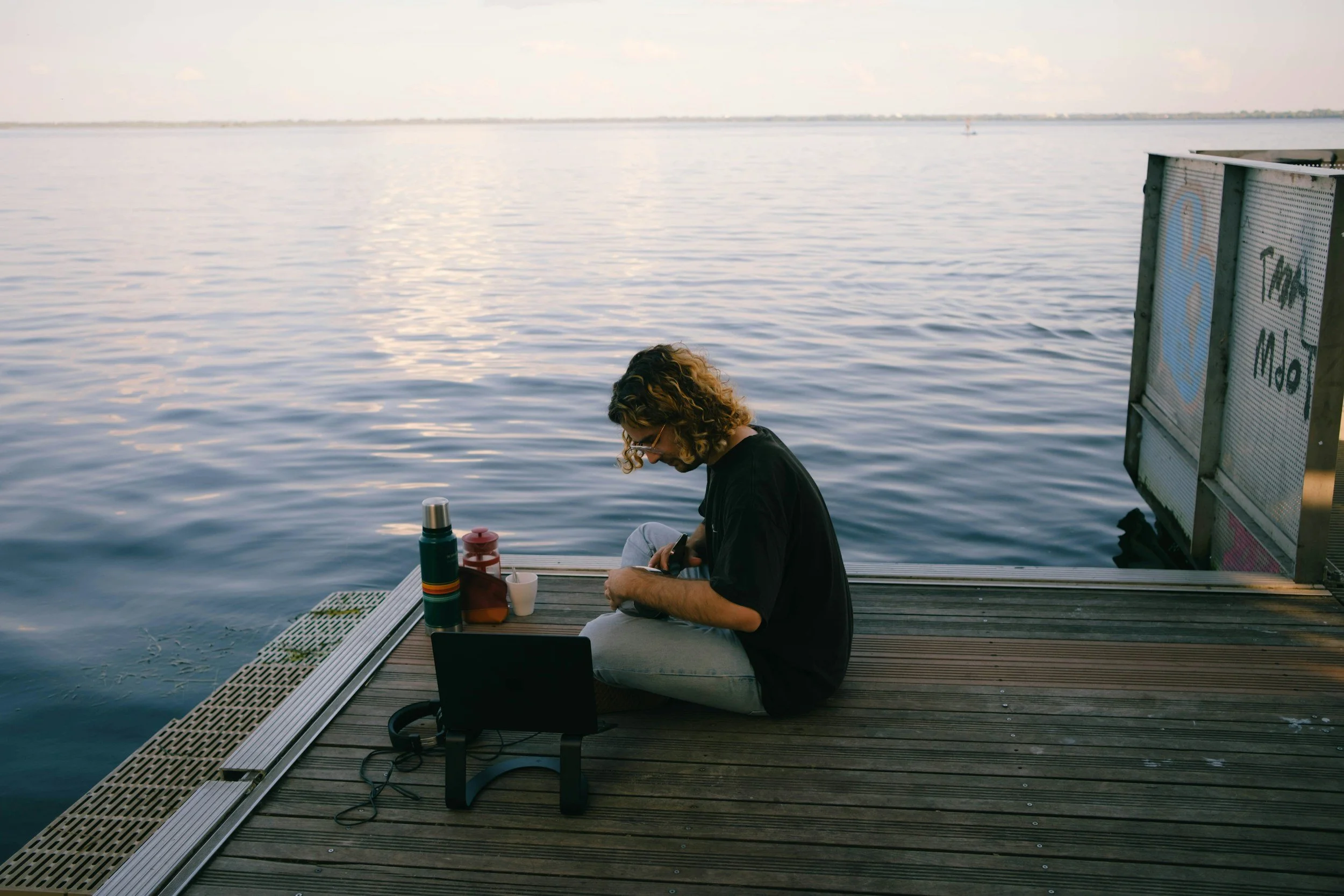 A person with curly hair and glasses sits on a dock by a body of water, looking at their phone. There are thermoses and cups on the dock, and a sign in the background that says 'Type a Child'.