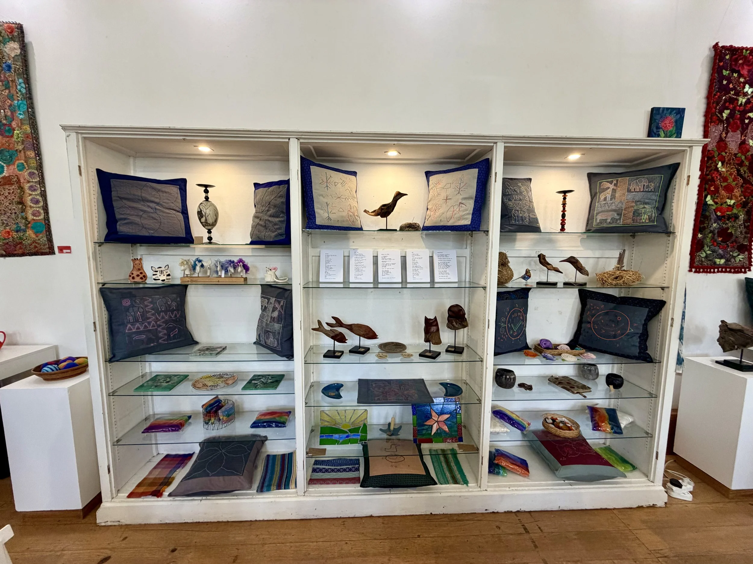 Display of handcrafted items on white shelving unit, including pillows, wood bird sculptures, and stained glass artwork, in an art gallery or shop setting.