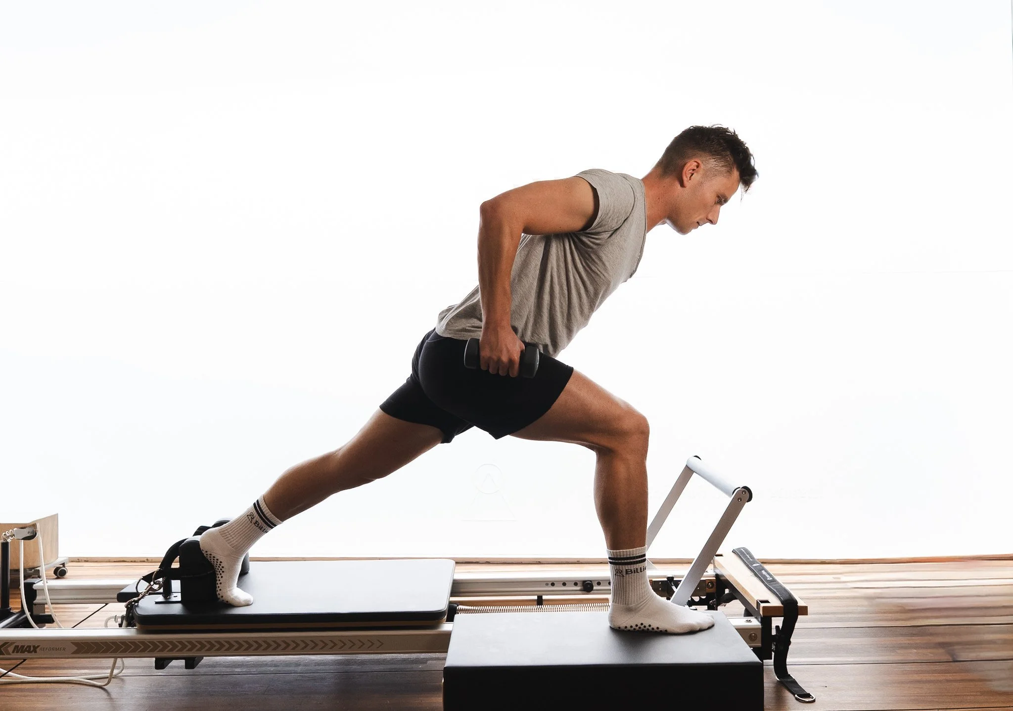 A young man in athletic clothing is performing a workout on a Pilates reformer machine, positioned with one foot on the platform and the other on the footrest, using hand weights, in a bright, minimal studio.
