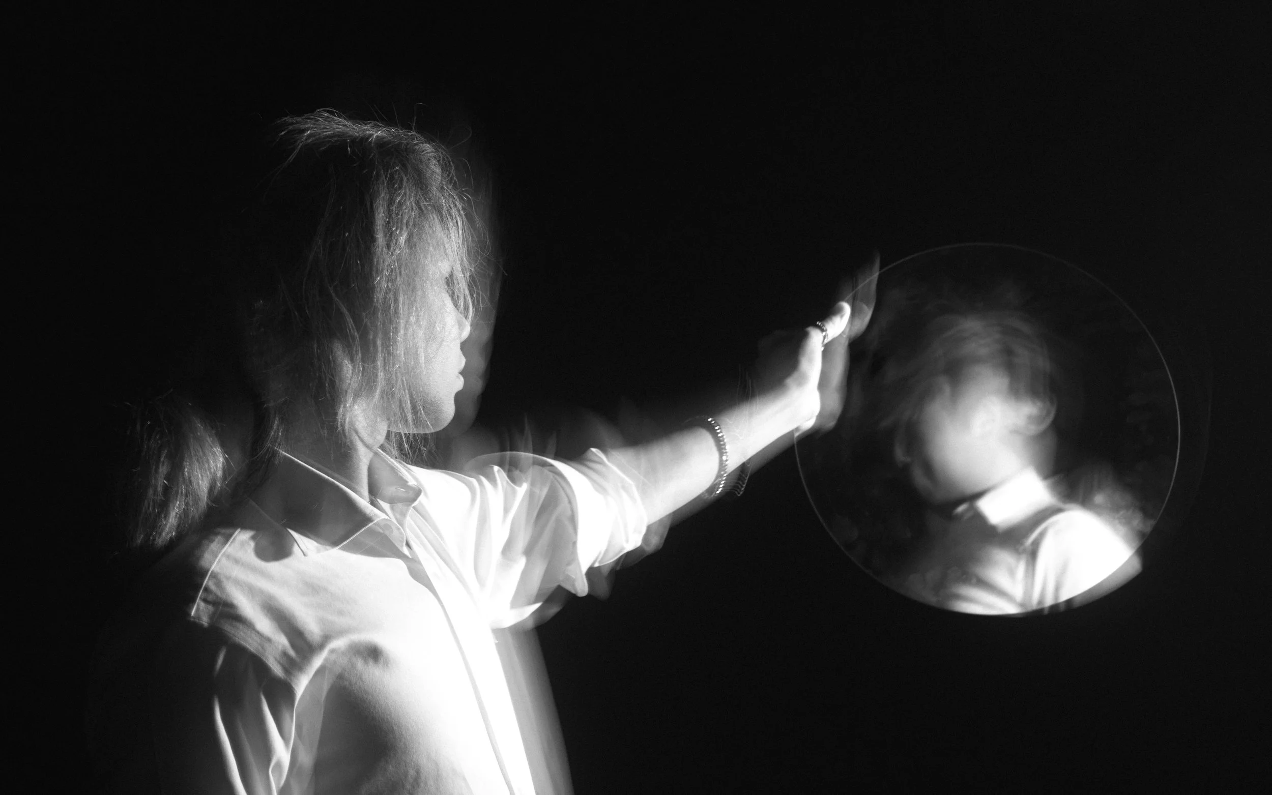 A black and white photo of a woman with curly hair in a white shirt, reaching out with her hand towards a blurred figure.