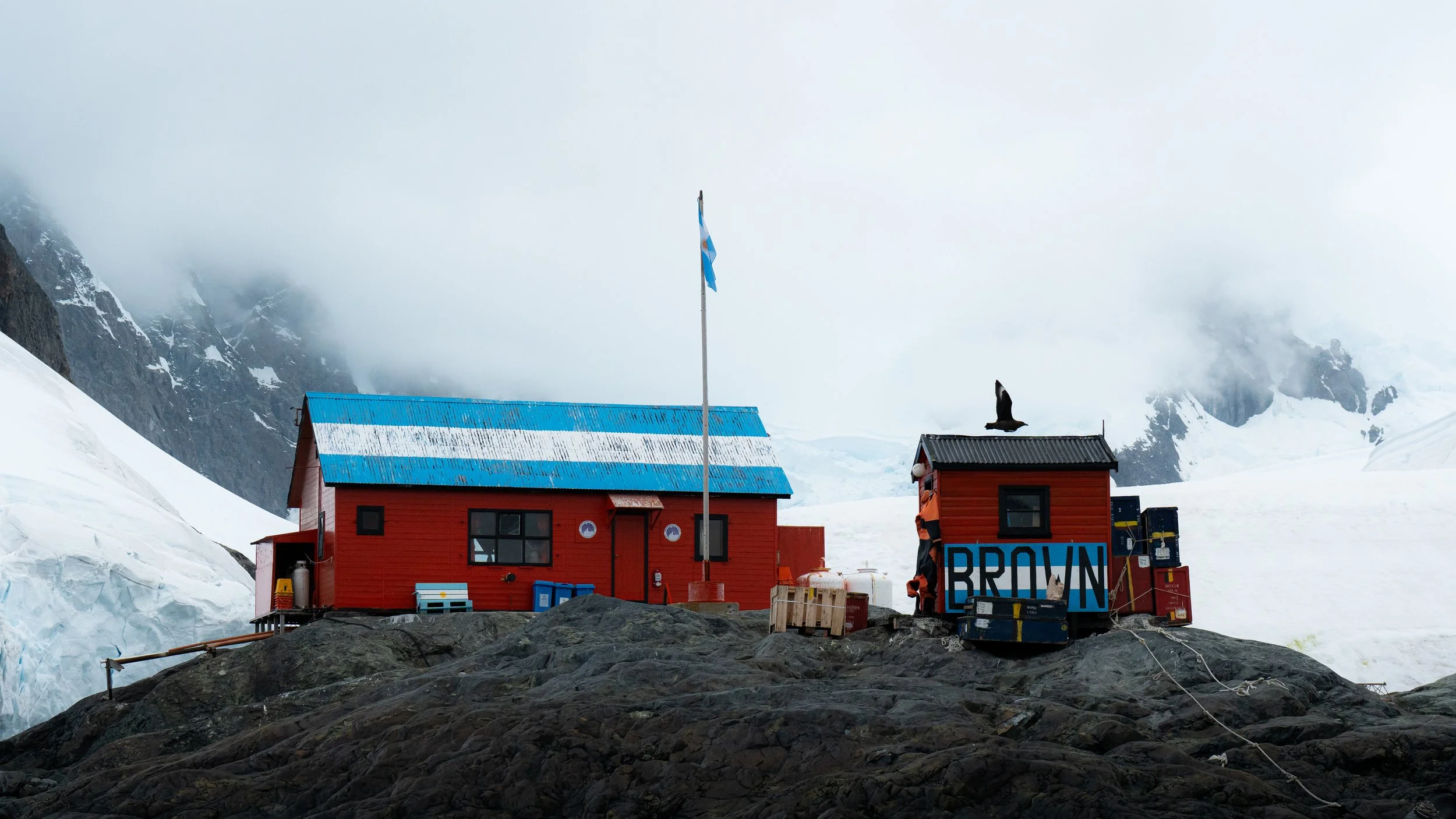 A small red building with a blue roof in a snowy mountain landscape. A tall flagpole with a blue and white flag. A bird is perched on the roof. Equipment and a sign that says 'BROWN' are in front of the building.
