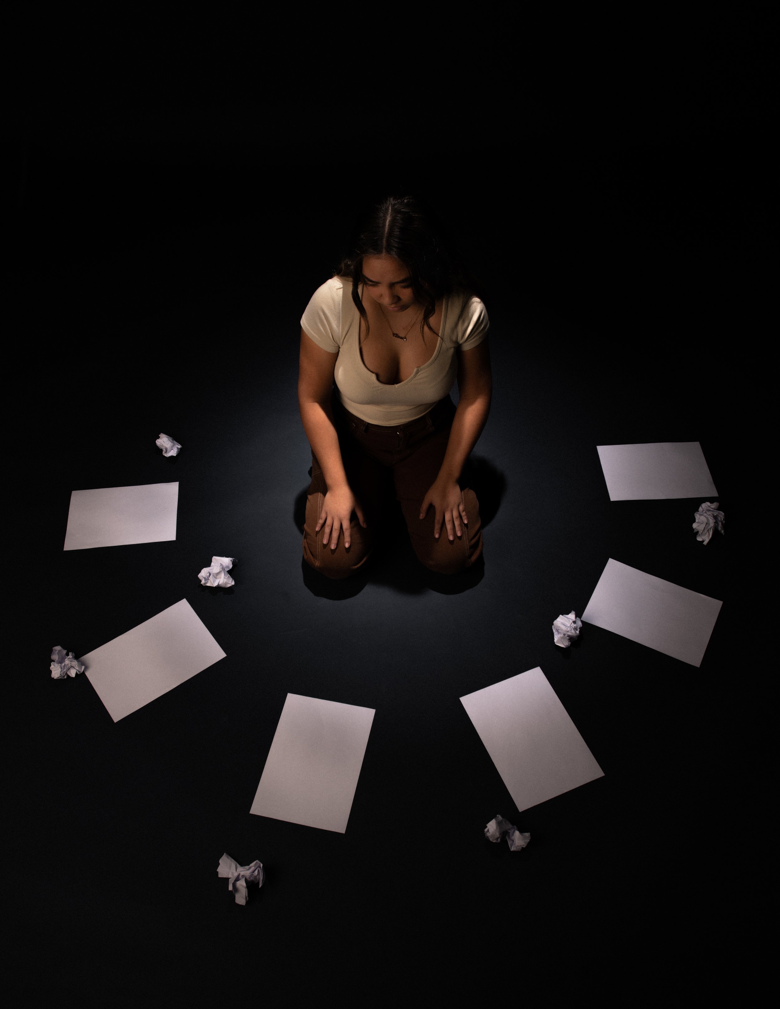 Woman kneeling on a dark floor surrounded by crumpled pieces of paper and blank sheets of paper.