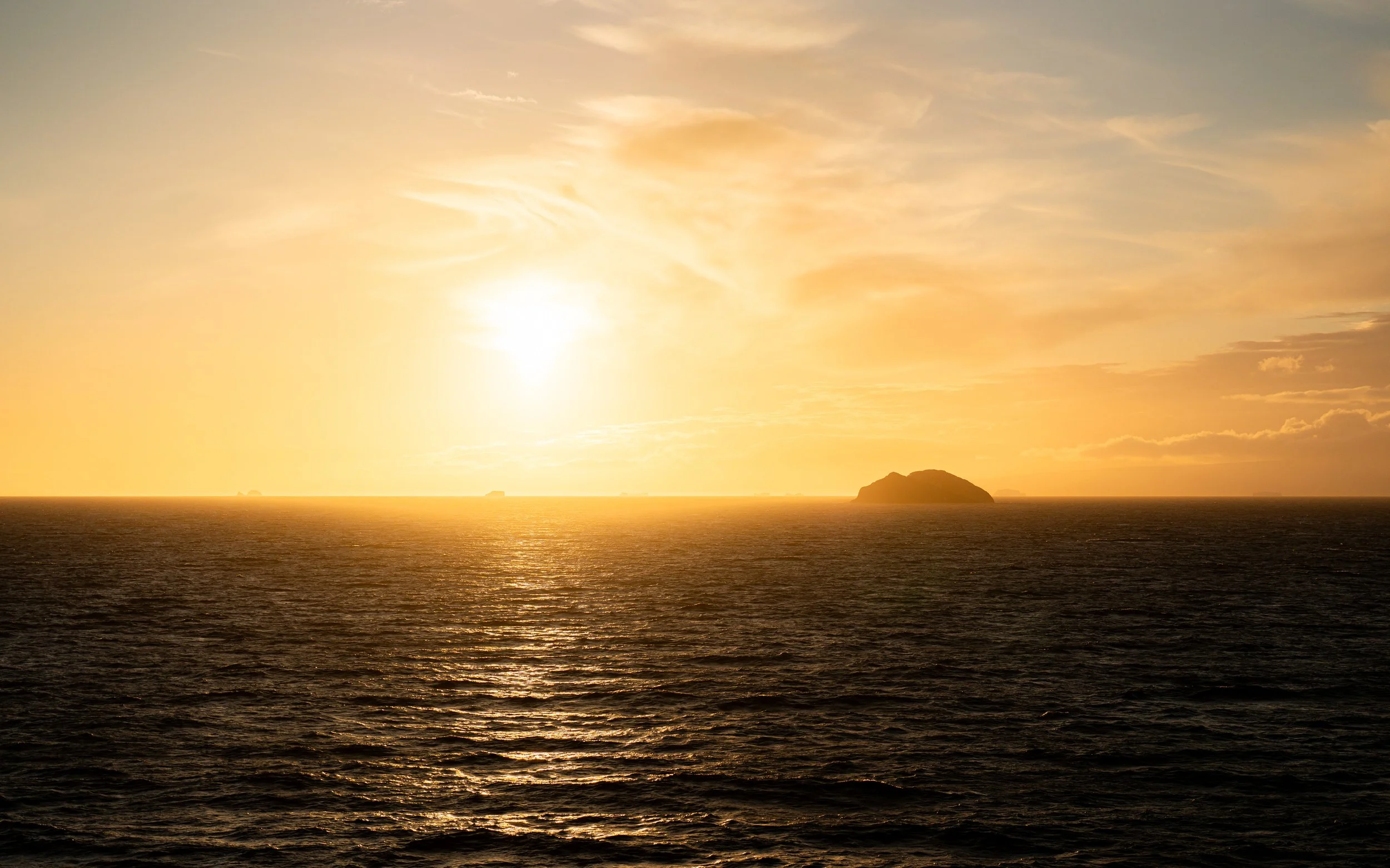 A sunset over the ocean with a small island silhouette on the horizon, sky filled with clouds and warm orange and yellow hues.