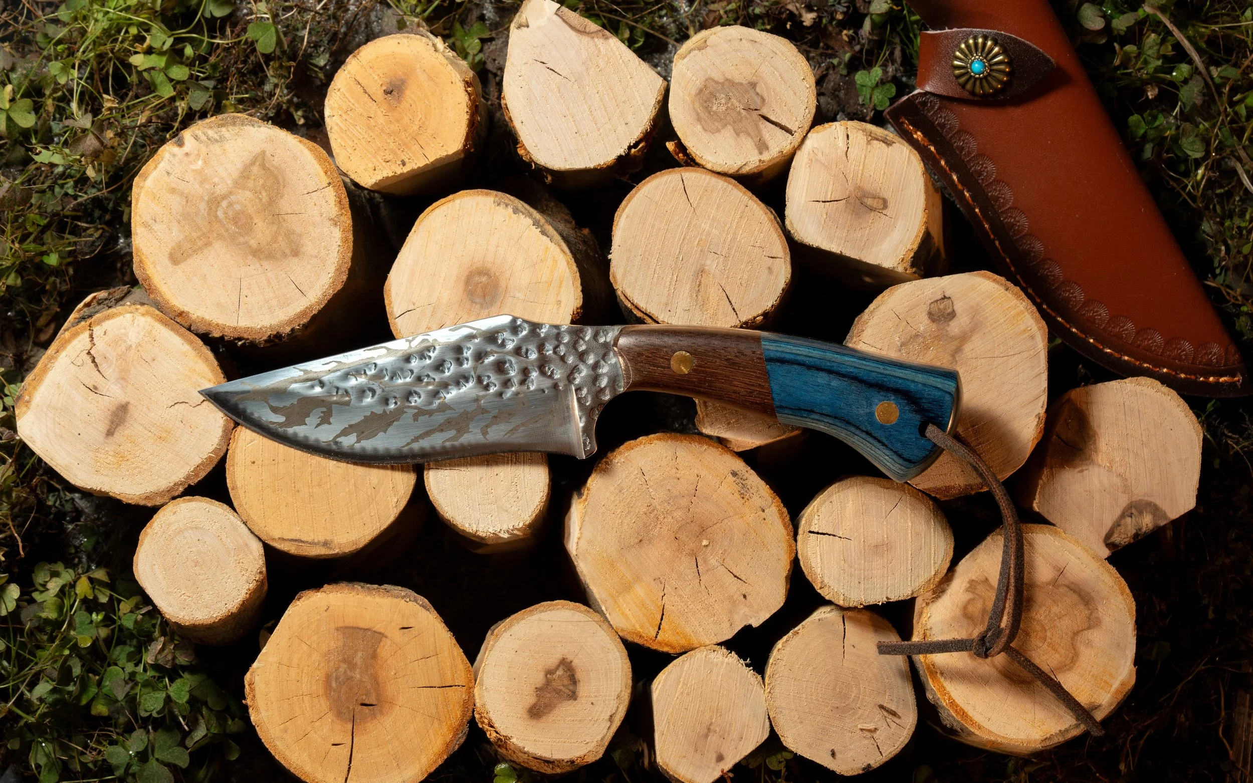 Stacked firewood logs placed on the ground, with a folding knife and a leather sheath lying on top.