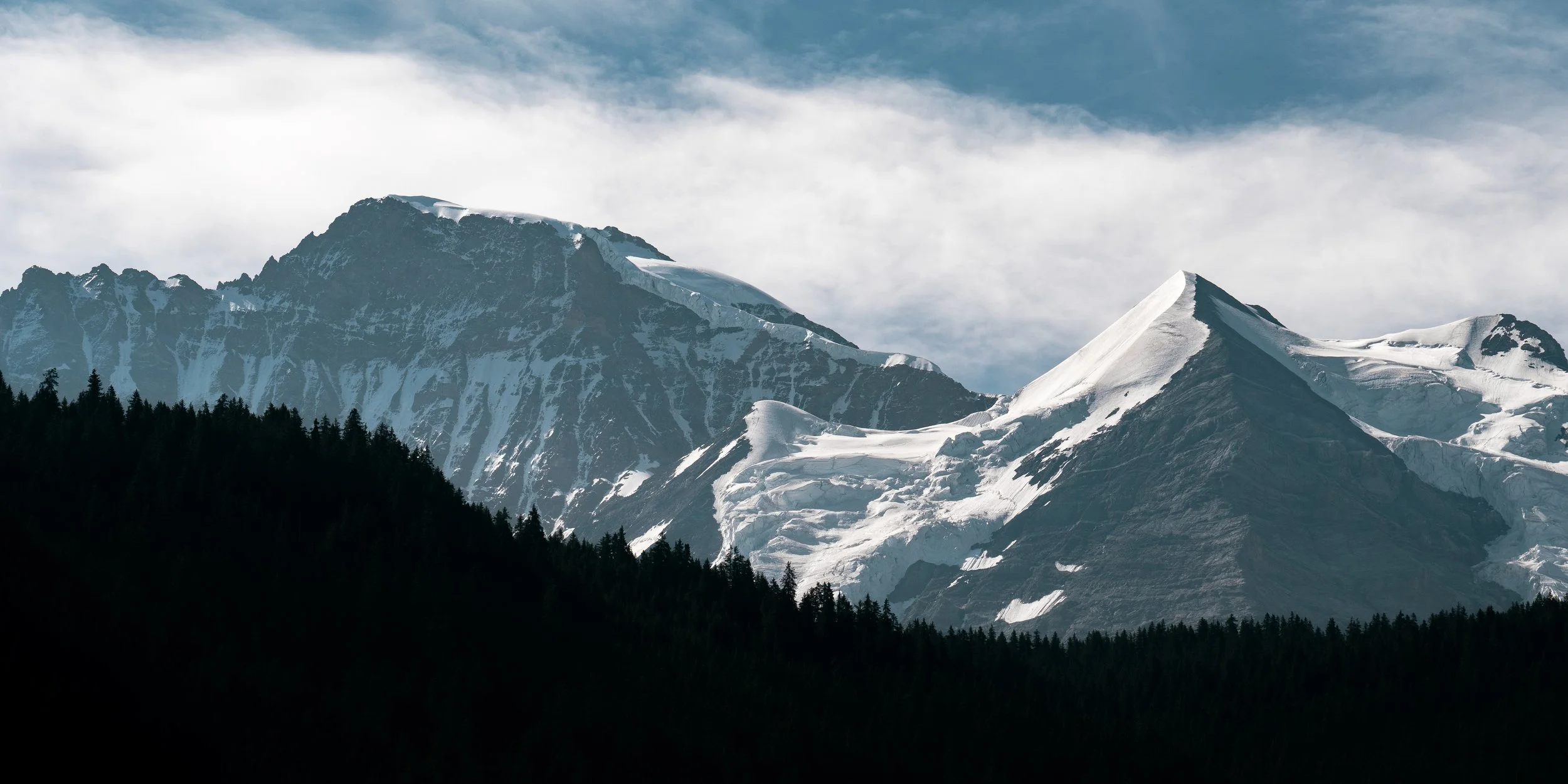 Snow-covered mountain peaks with a forest in the foreground under a partly cloudy sky.